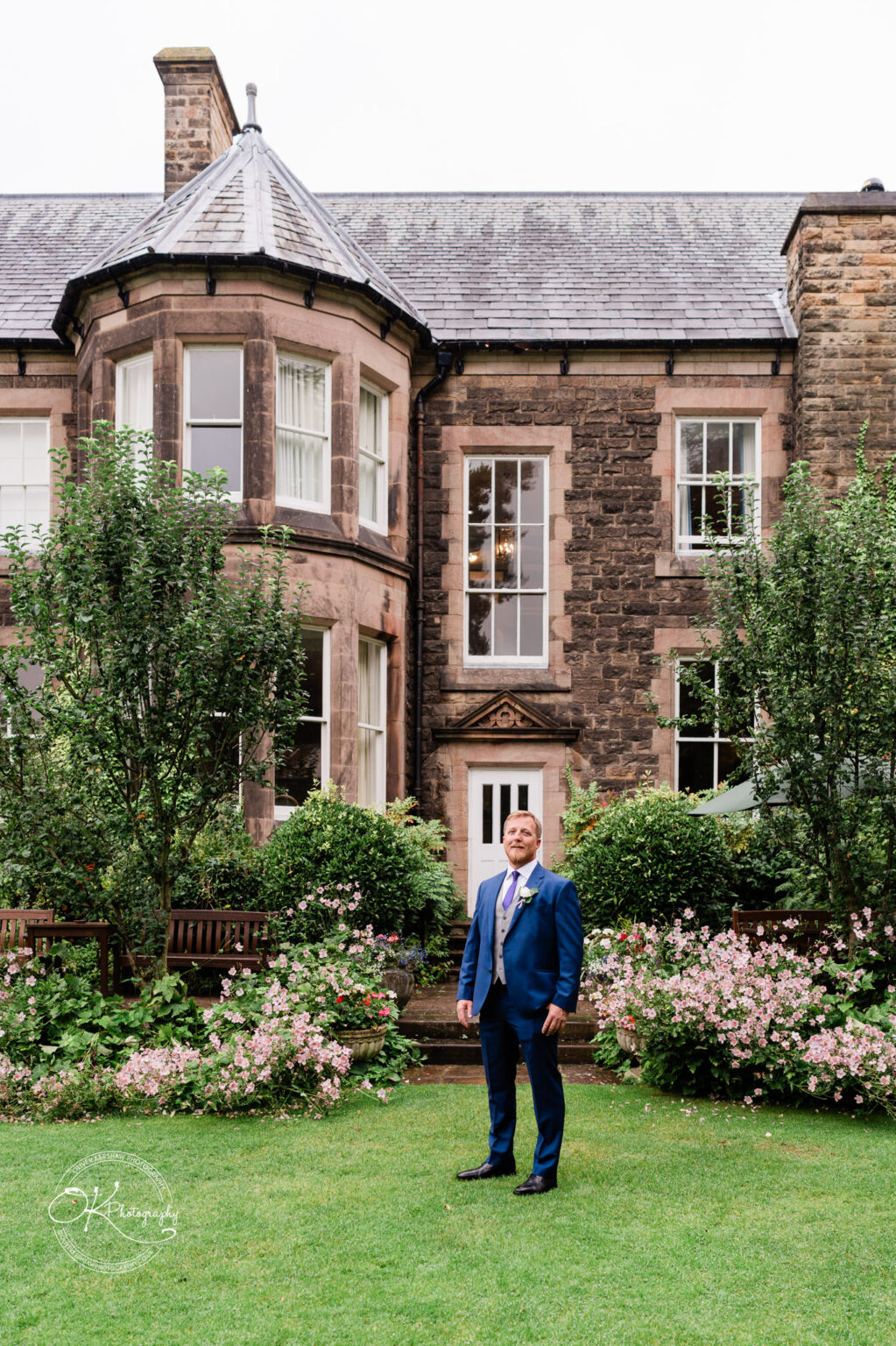 A man in a blue suit stands in front of the historic Makeney Hall Hotel, surrounded by lush gardens and blooming flowers.