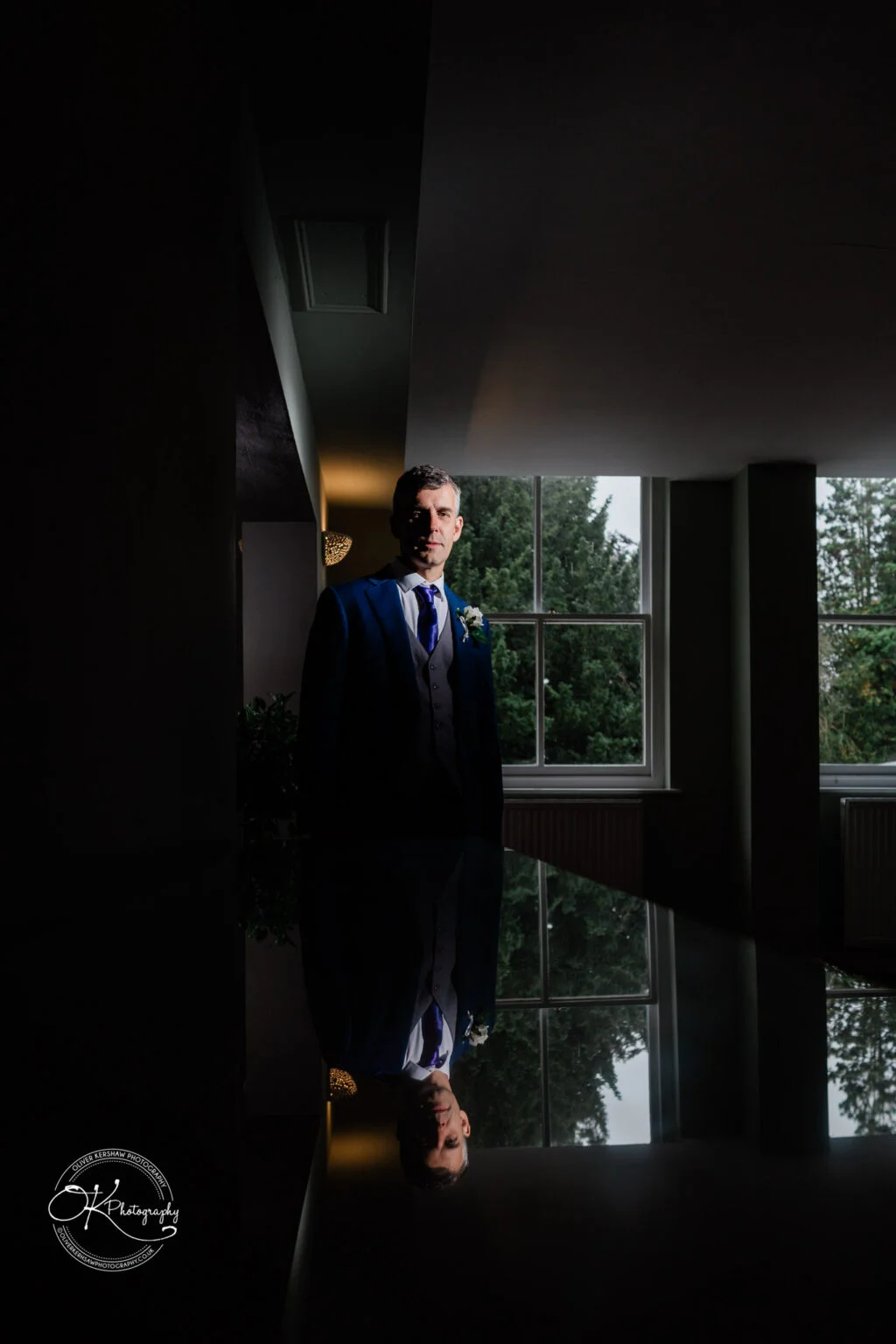 Makeney Hall Hotel - Wedding Photography A man in a suit standing indoors near a window, with a reflection of himself on a polished surface.