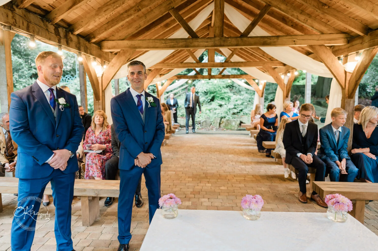 Makeney Hall Hotel - Wedding Photography Two men in blue suits stand under a wooden gazebo decorated with draped white fabric, while seated guests in formal attire await the start of a ceremony.