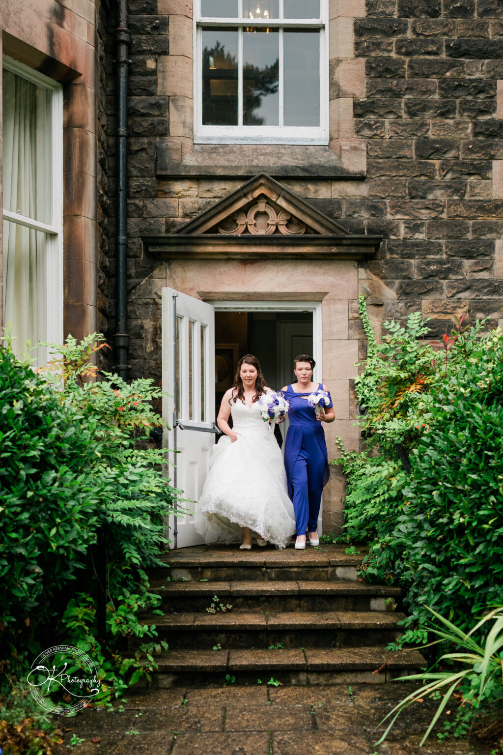 Two women, one in a white wedding dress and the other in a blue outfit, walking down steps outside a stone building with greenery around.