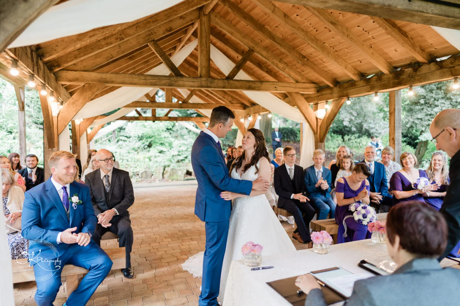 Makeney Hall Hotel - Wedding Photography A bride and groom stand facing each other under a wooden pavilion adorned with white drapery at Makeney Hall Hotel, witnessed by seated guests.