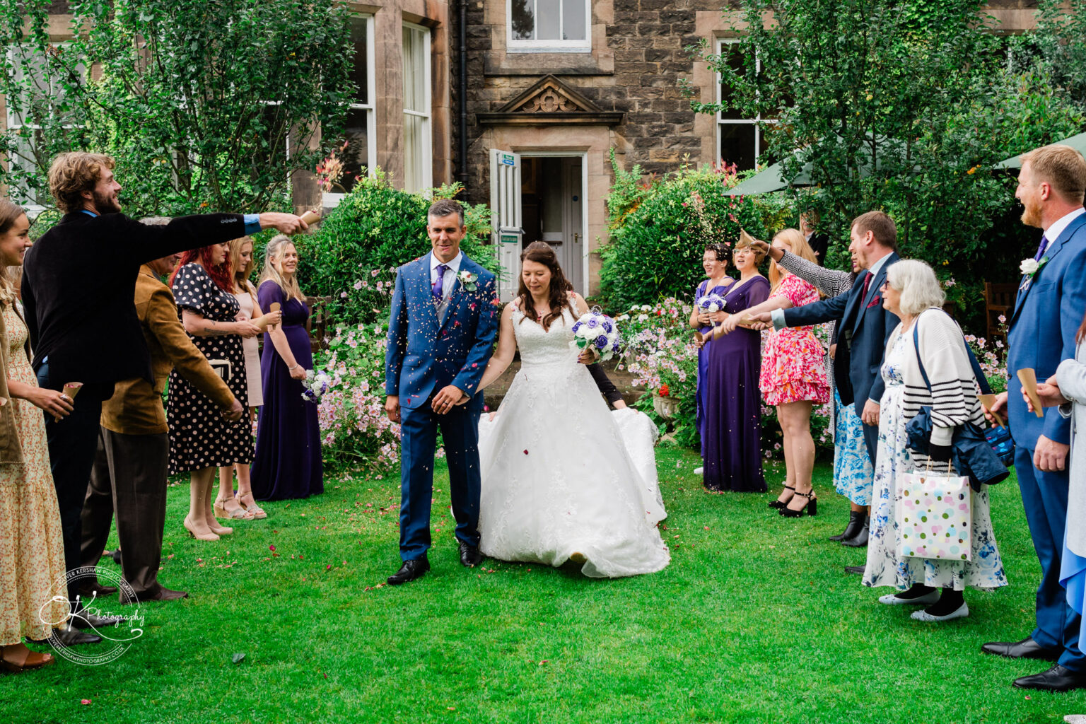 A newlywed couple walks through a garden amid a shower of confetti, surrounded by smiling guests, in front of a historic building.