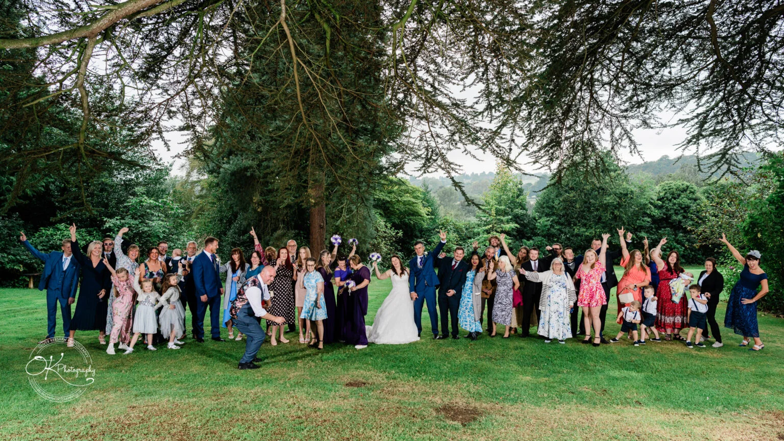Makeney Hall Hotel - Wedding Photography A large group posing for a wedding photo on a lawn with trees in the background.