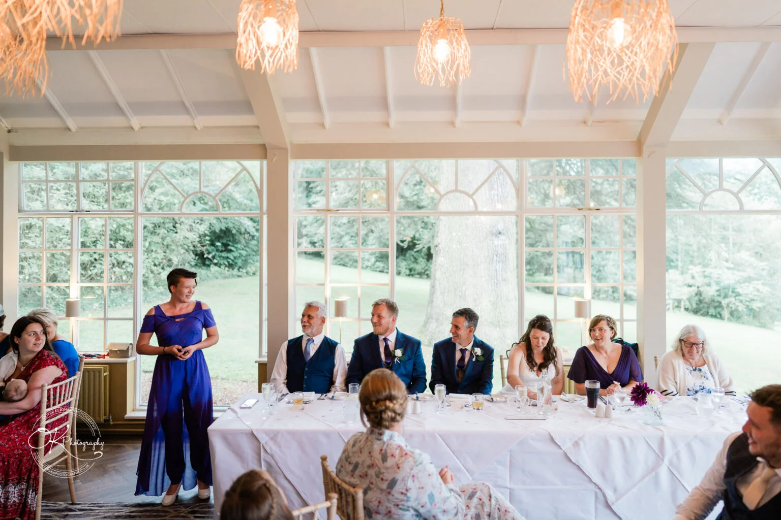 Makeney Hall Hotel - Wedding Photography A wedding speech is being given at a long table decorated with white tablecloths and set with glasses and plates, in a room with large windows at Makeney Hall Hotel.