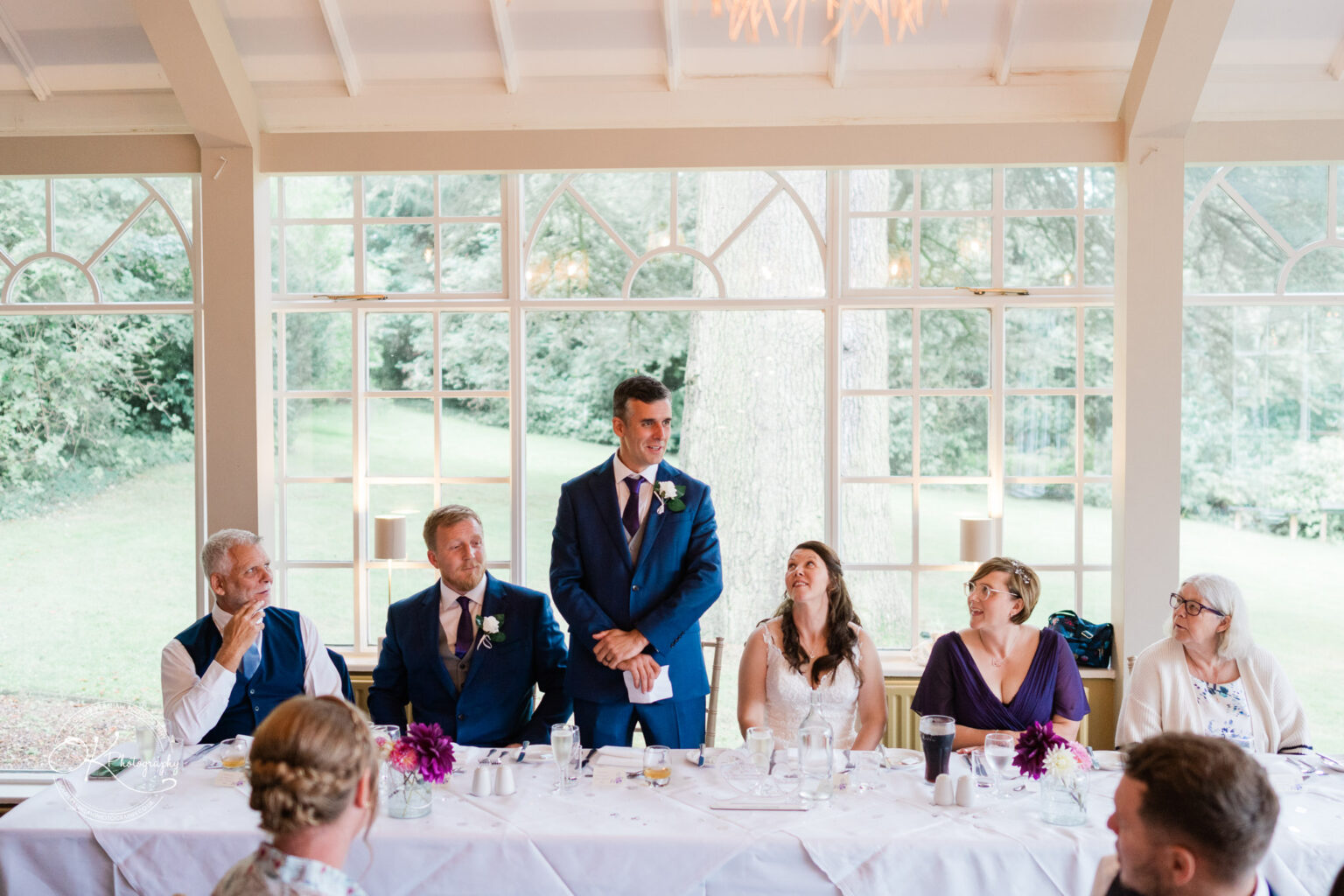 A wedding reception at Makeney Hall Hotel with a man in a blue suit giving a speech, seated guests flanking him at a long table, and large windows showcasing greenery in the background.