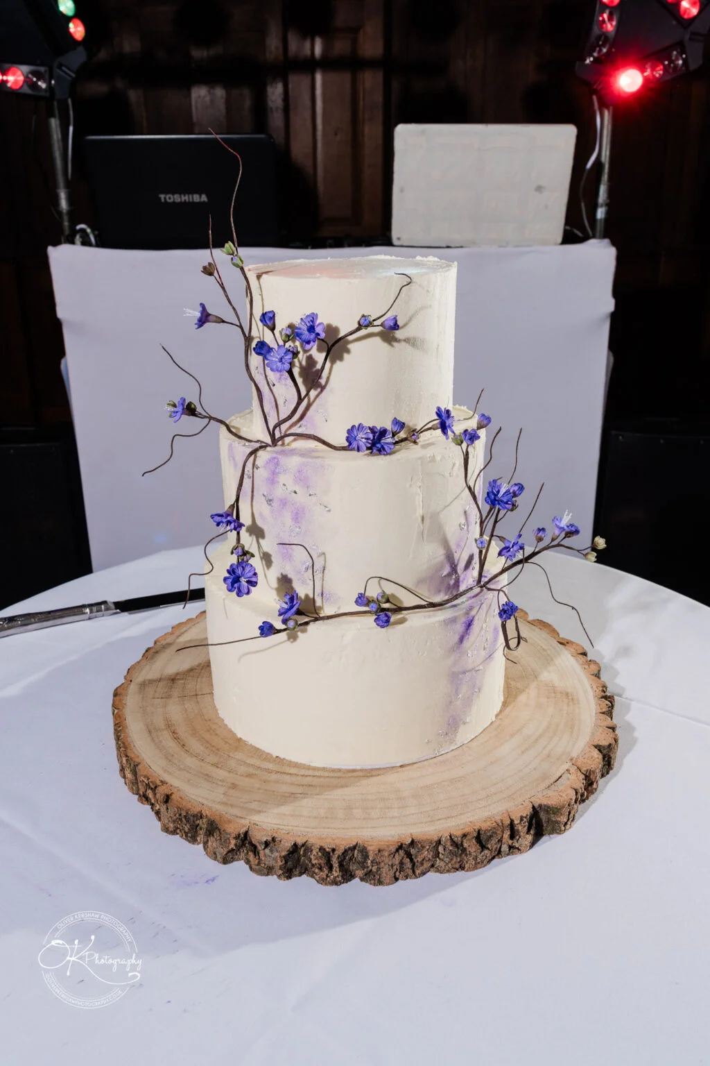 Makeney Hall Hotel - Wedding Photography Three-tiered wedding cake with white frosting and decorated with delicate purple flowers, placed on a wooden slab base.