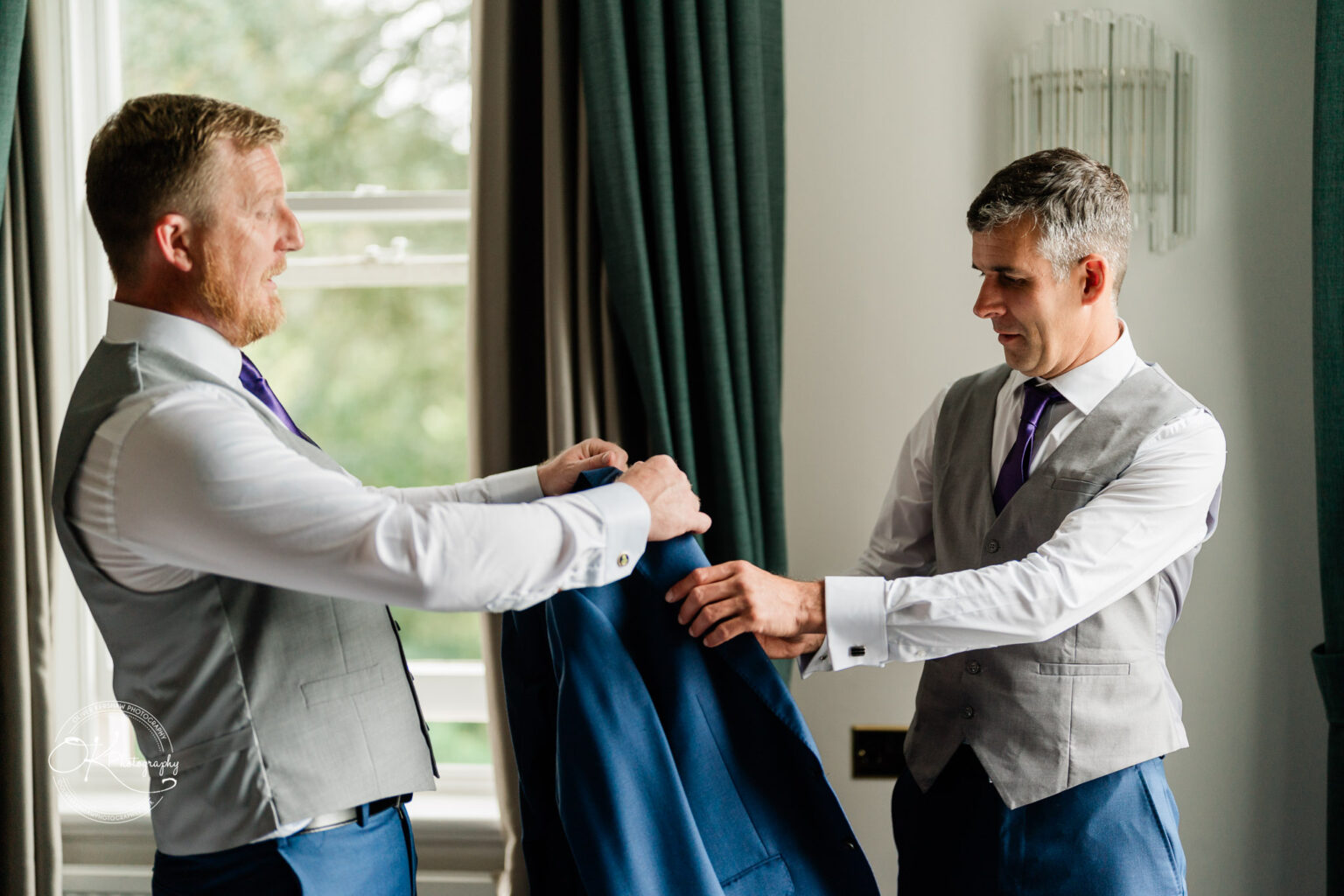 Two men in grey vests and purple ties preparing a blue suit jacket, standing by a window with green curtains at Makeney Hall Hotel.