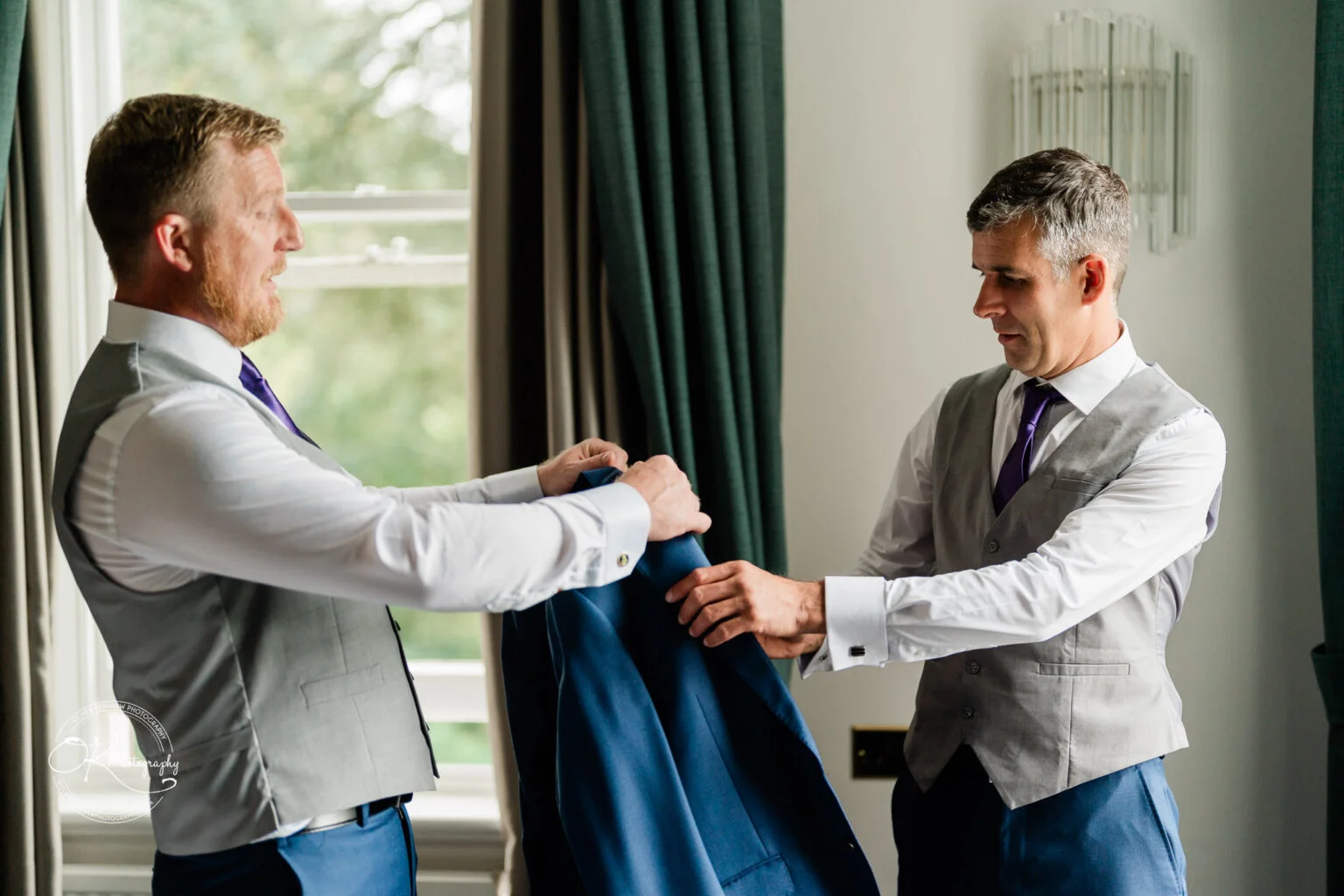 Makeney Hall Hotel - Wedding Photography Two men in grey vests and purple ties preparing a blue suit jacket, standing by a window with green curtains at Makeney Hall Hotel.