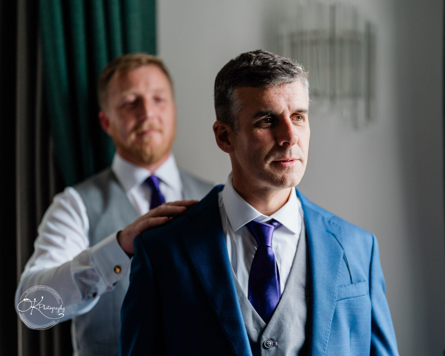 Two men in formal attire, one adjusting the other's suit jacket in a well-lit room.