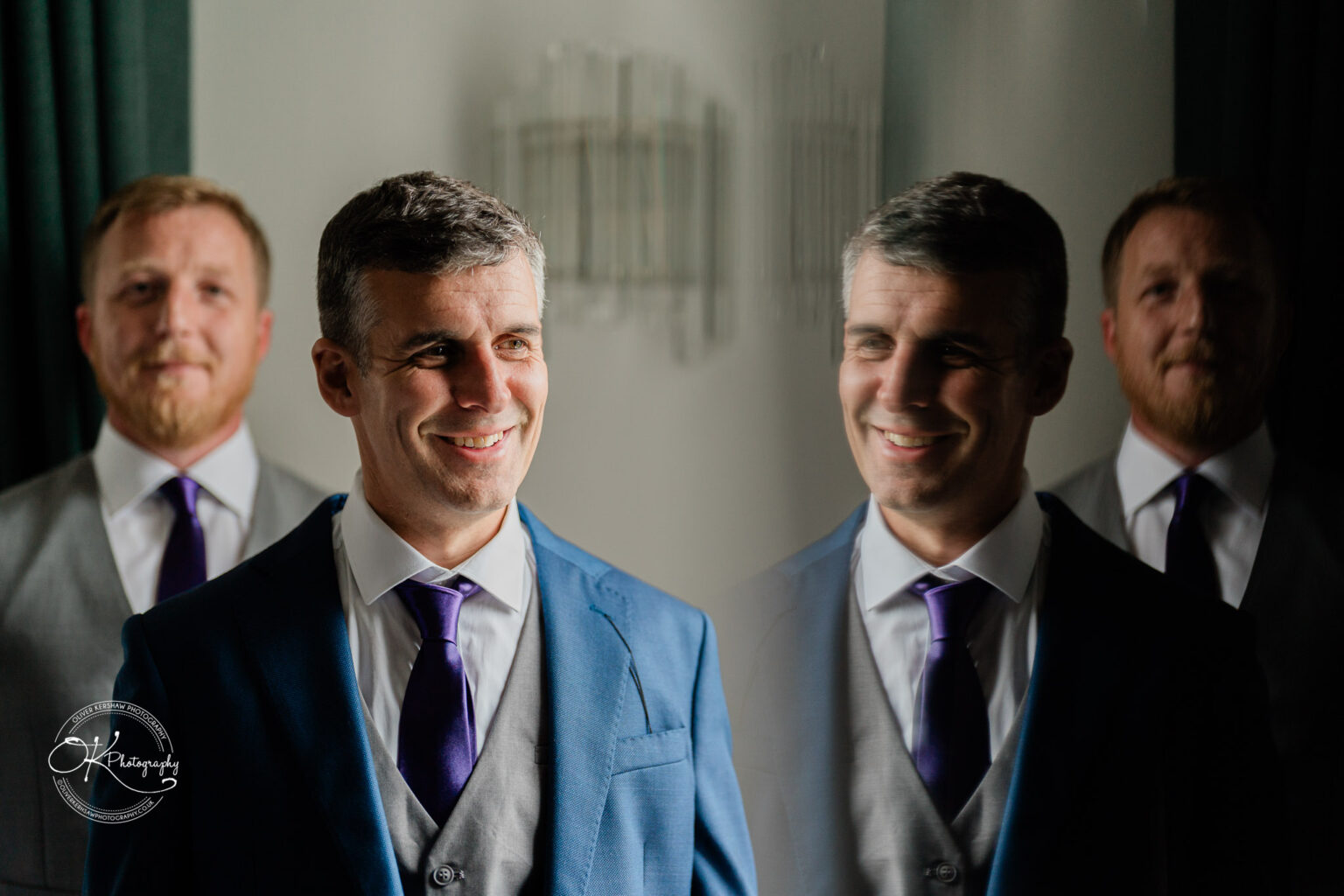 Two men dressed in suits and ties, one in the foreground smiling with his reflection visible, and another in the background, standing inside a room at Makeney Hall Hotel.
