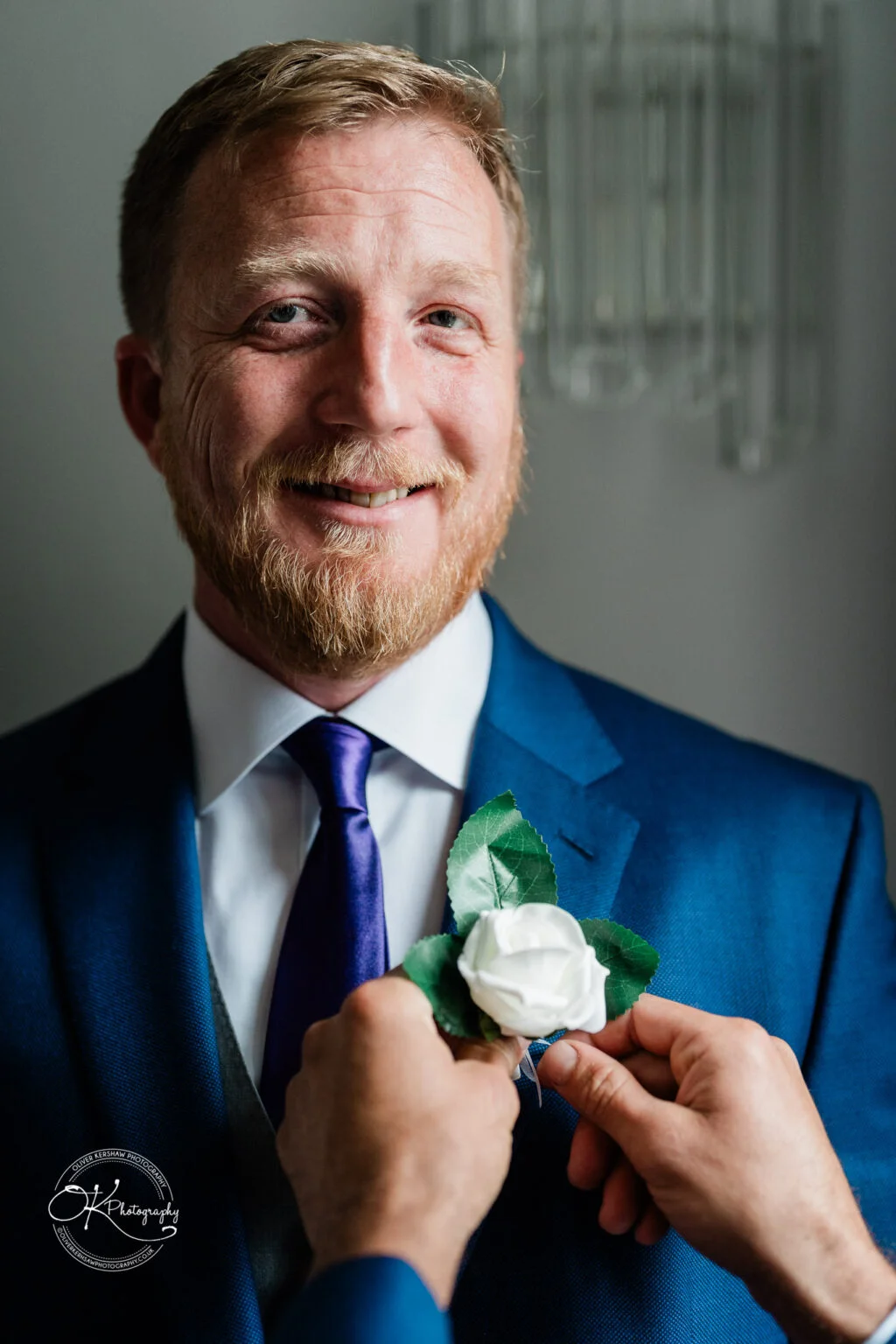 Makeney Hall Hotel - Wedding Photography A smiling man in a blue suit with a white shirt and purple tie, having a white rose boutonnière pinned to his lapel by another person.