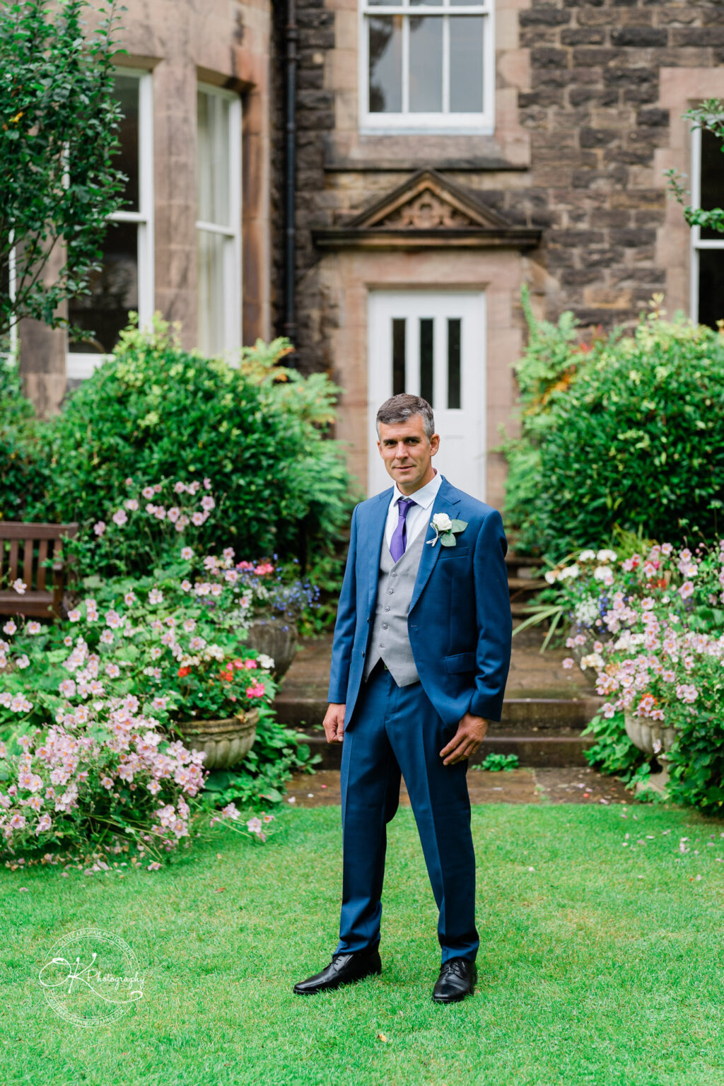 A man in a blue suit and tie with a white boutonnière stands on a lawn in front of Makeney Hall Hotel, surrounded by greenery and flowers.
