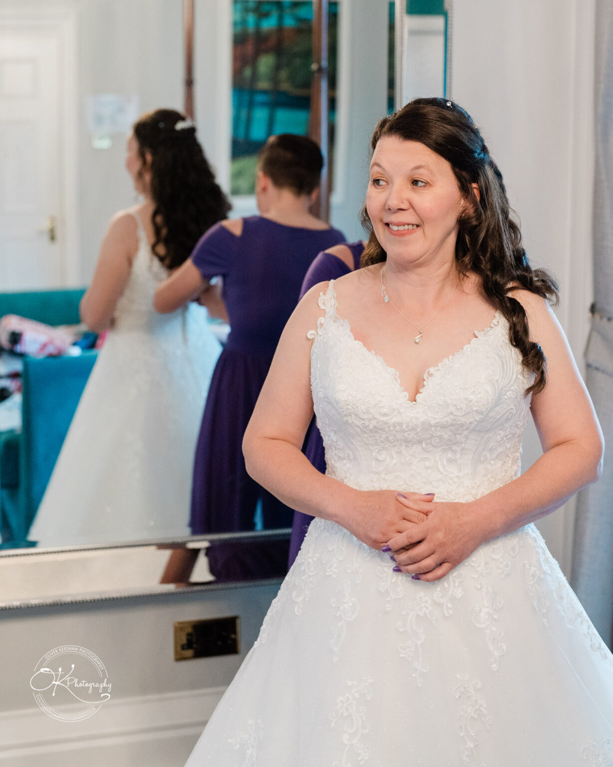 A bride in a white wedding dress and a bridesmaid in a purple dress stand in front of a mirror in a room.