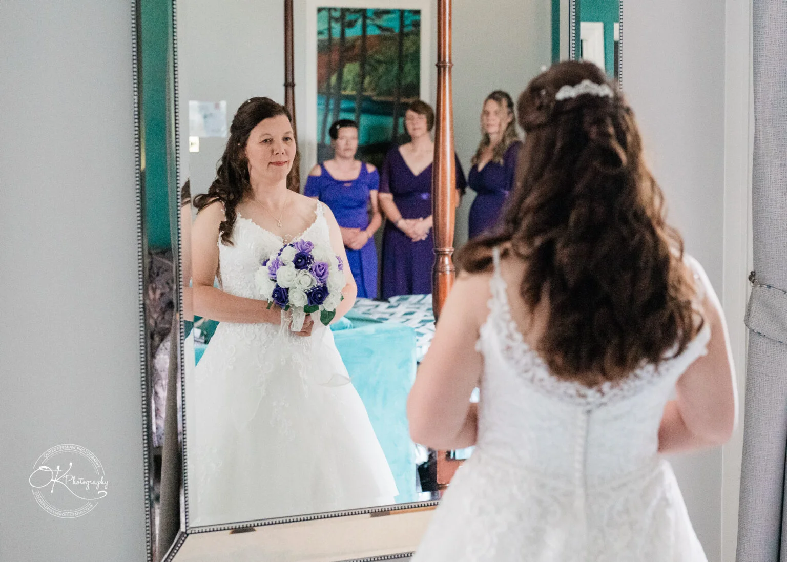 Makeney Hall Hotel - Wedding Photography A bride in a white gown holds a bouquet of purple and white flowers, looking at her reflection in a mirror, with bridesmaids in purple dresses standing in the background.