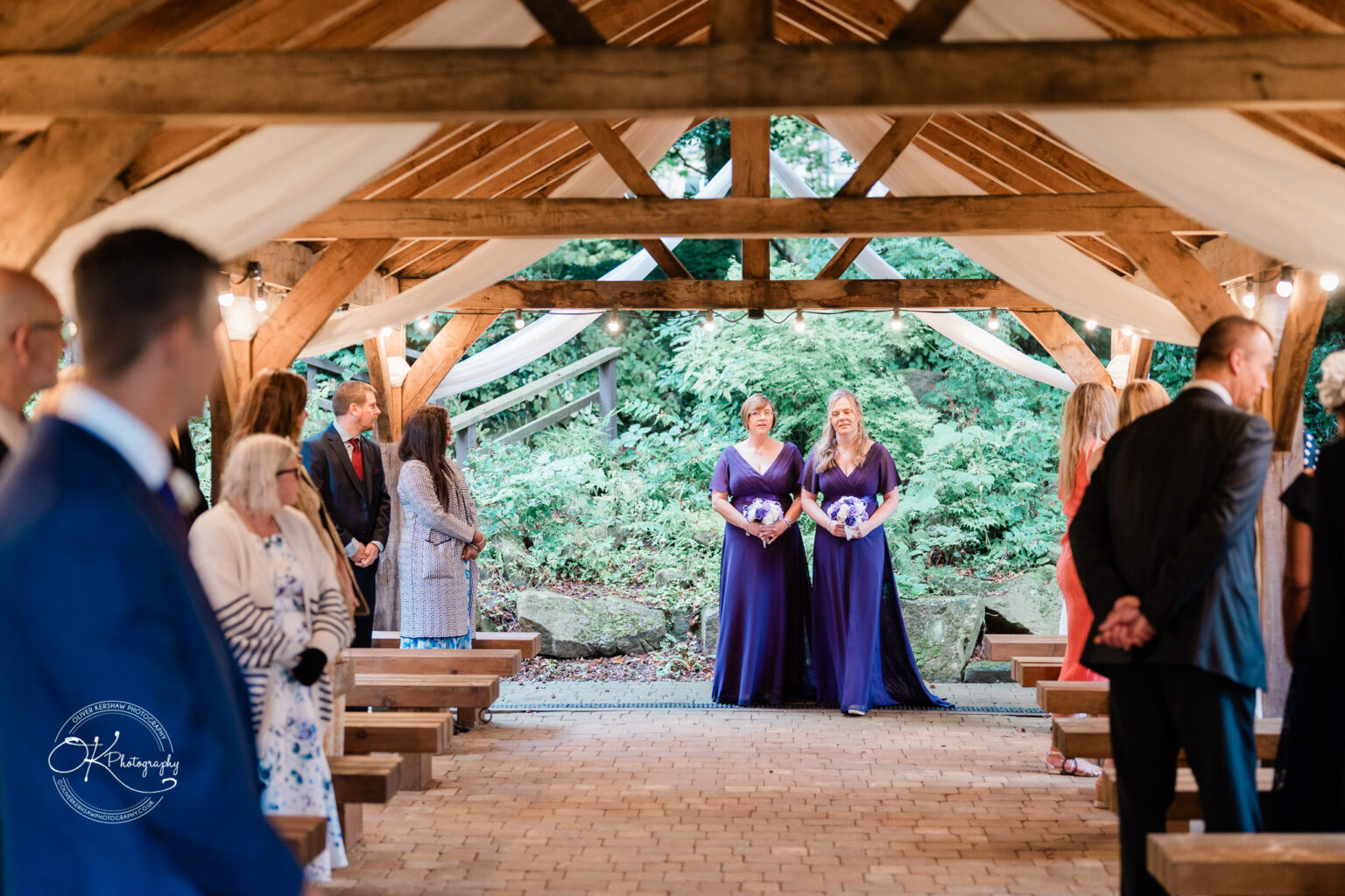 An outdoor wedding ceremony at Makeney Hall Hotel, with two bridesmaids in purple dresses holding bouquets, and guests standing on either side of the aisle under a wooden gazebo decorated with white fabric.