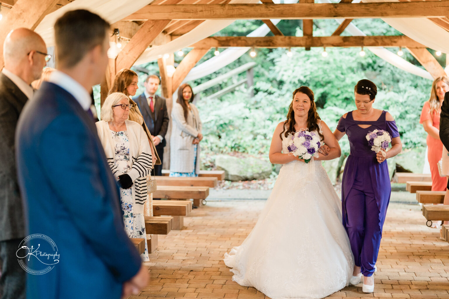 A bride in a white wedding dress walks down the aisle with a bridesmaid in a purple dress, holding bouquets of purple and white flowers, in an outdoor ceremony at a wooden venue with guests watching.
