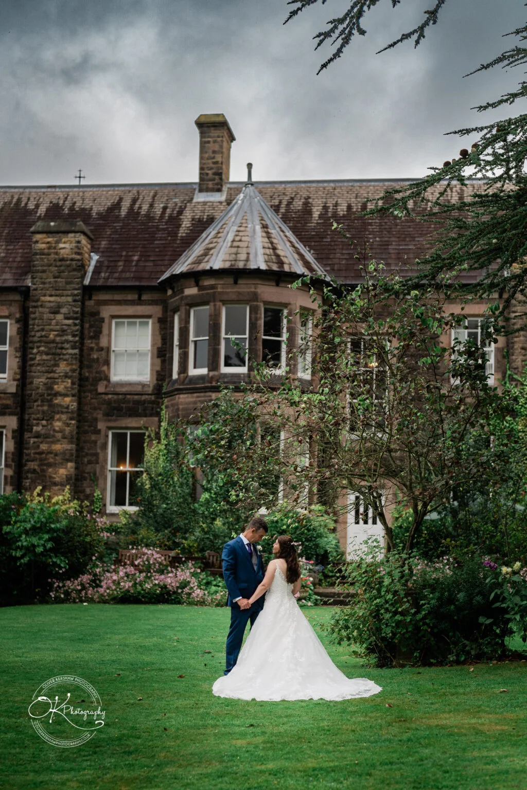 Makeney Hall Hotel - Wedding Photography A couple in wedding attire poses on a lawn in front of Makeney Hall Hotel, a historic stone building with a turret and lush gardens.
