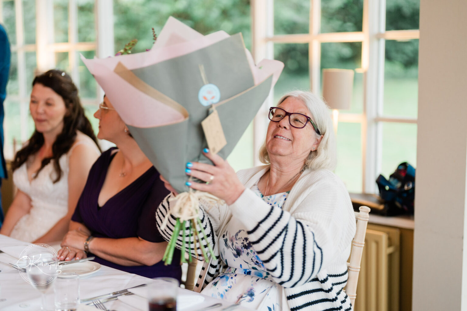 An elderly woman holding a bouquet of flowers, sitting at a table with two other women at Makeney Hall Hotel.
