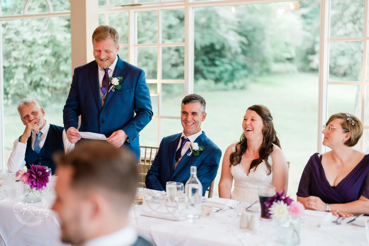 Makeney Hall Hotel - Wedding Photography A man gives a speech at a wedding reception with the bride, groom, and guests smiling at the head table.