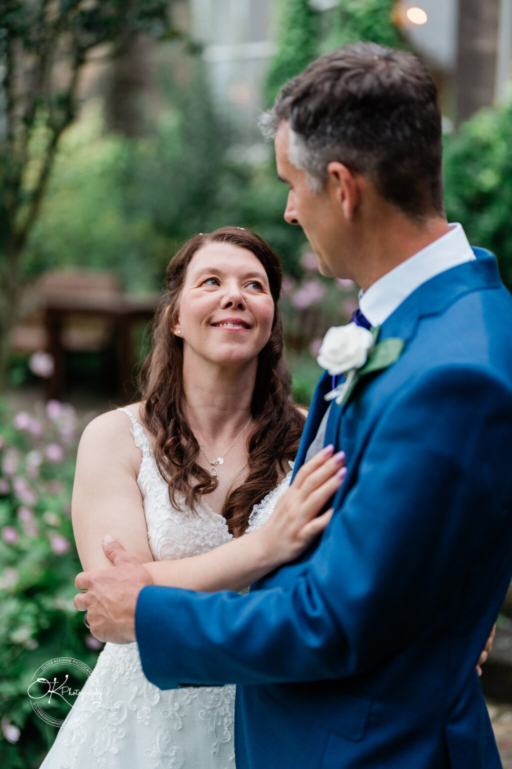 A couple embracing and looking at each other lovingly outdoors, with greenery and flowers in the background.