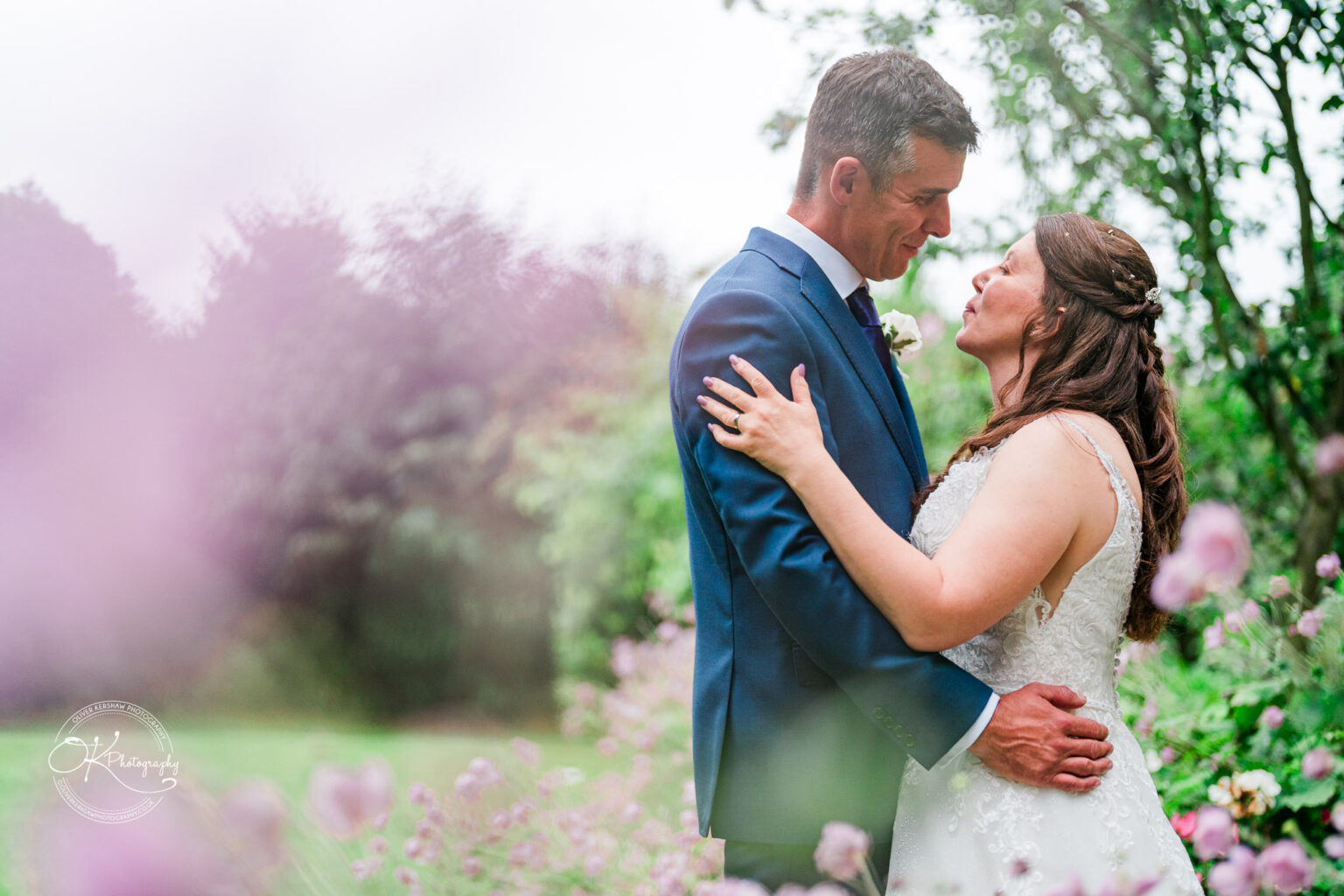 A bride and groom embracing in a garden setting, surrounded by pink flowers.