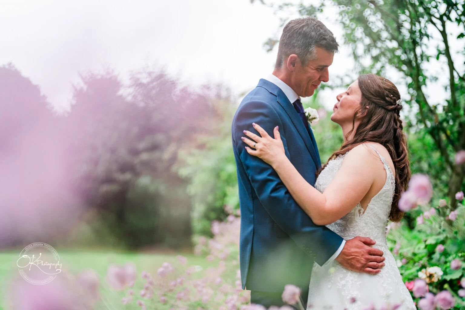 Makeney Hall Hotel - Wedding Photography-By-Oliver-Kershaw-Photography-OK906672 A bride and groom embracing in a garden setting, surrounded by pink flowers.