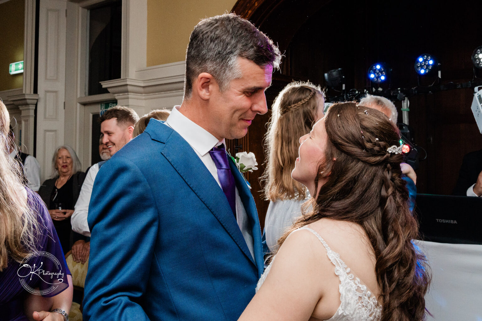A wedding couple dancing, looking into each other's eyes, surrounded by guests in a warmly-lit reception hall.