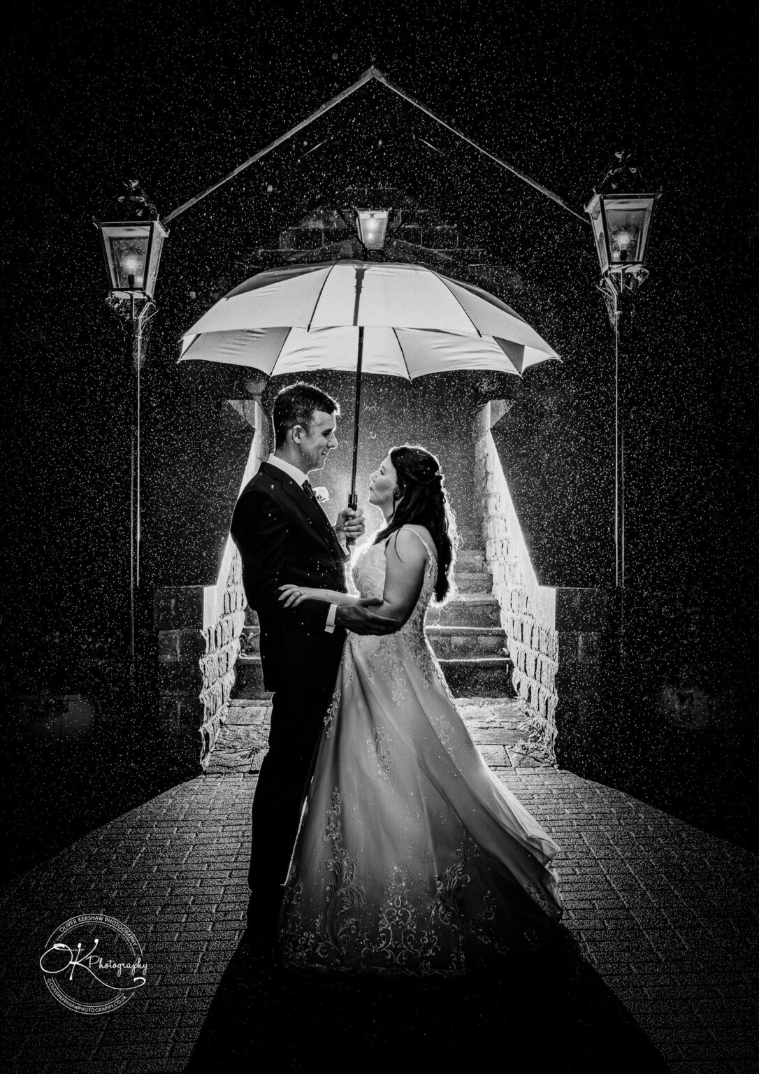 Makeney Hall Hotel - Wedding Photography Couple embracing under an umbrella at night in front of illuminated stairs with rain falling around them.