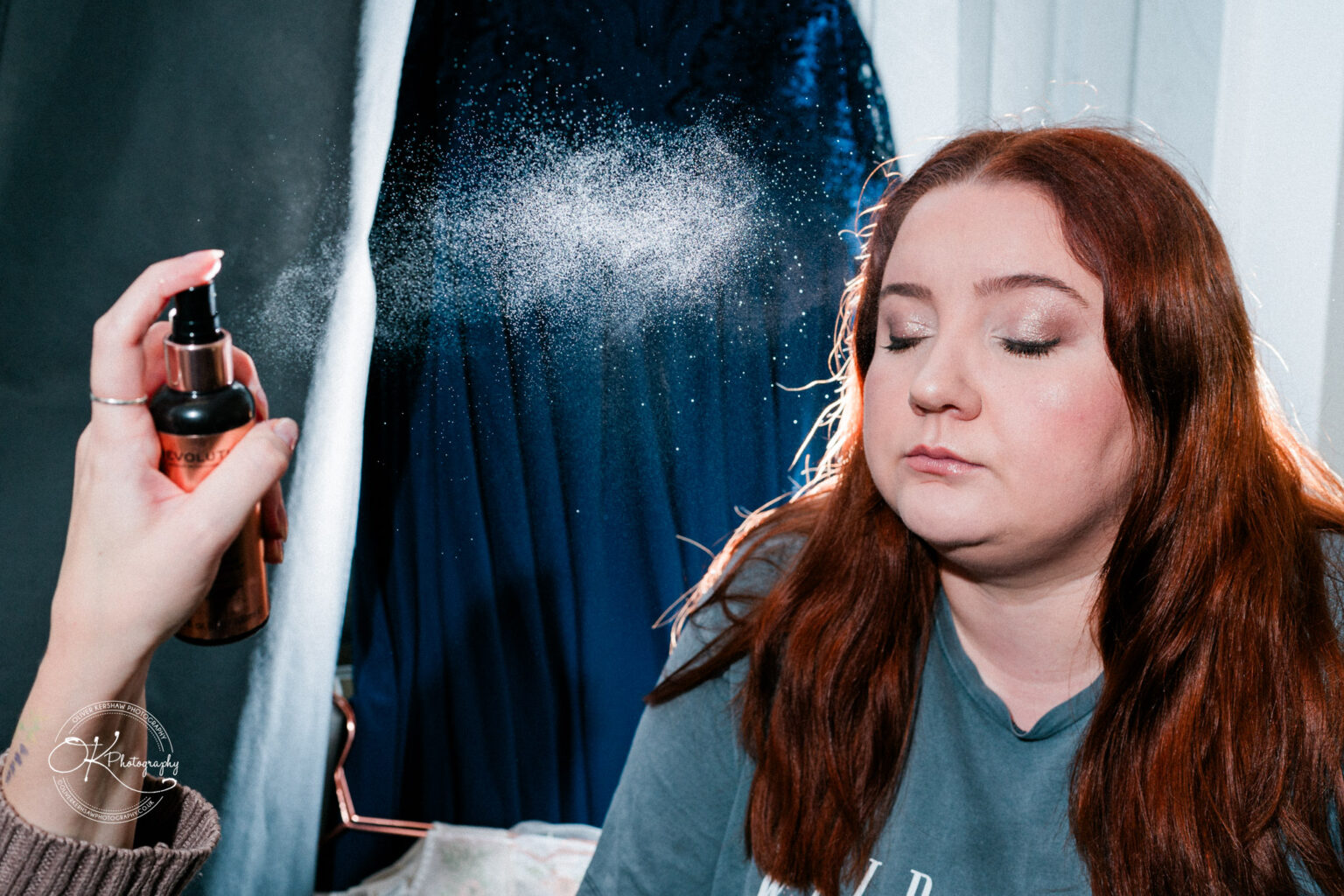 A person with long red hair and closed eyes being sprayed with setting spray during a makeup application.