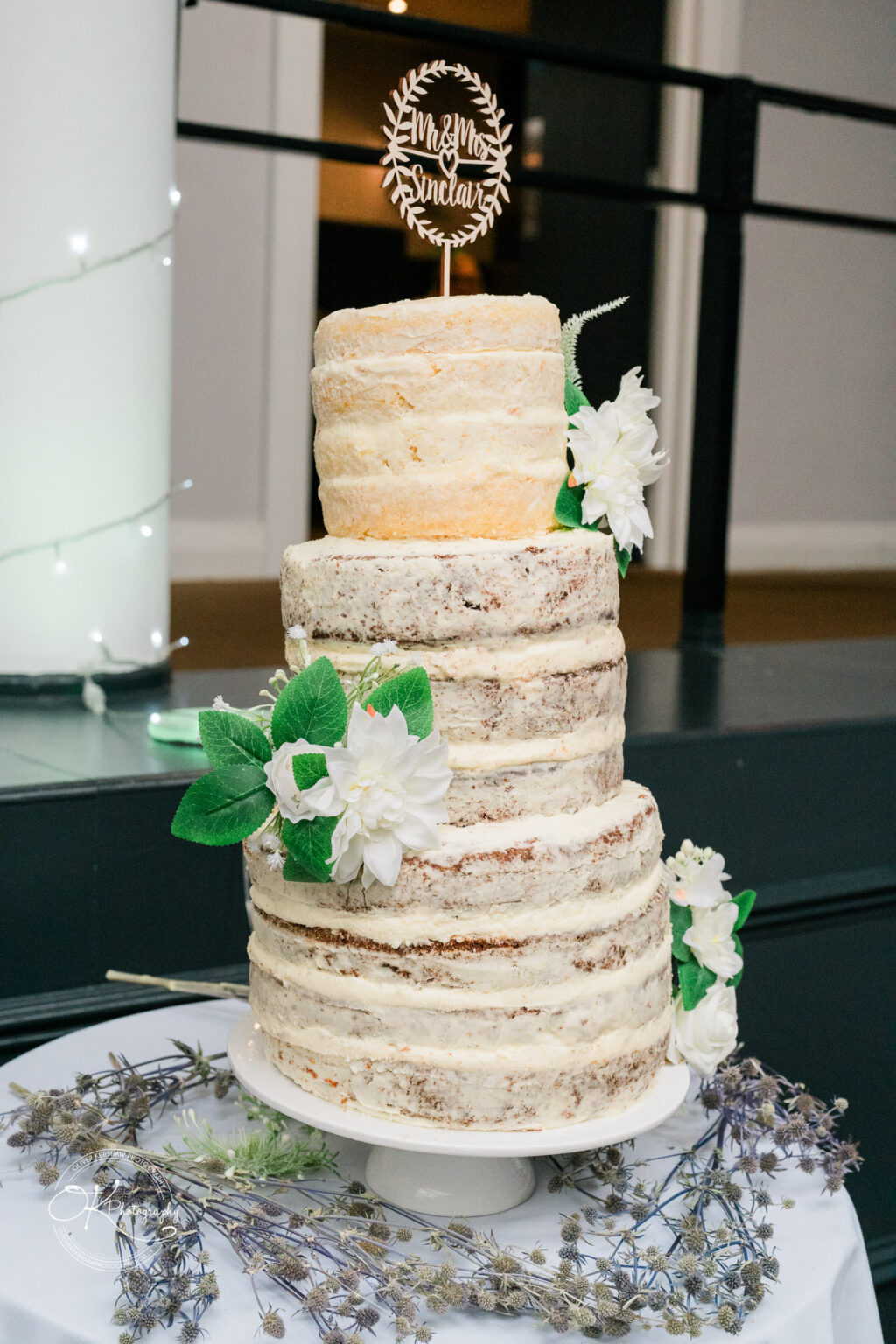 Three-tiered semi-naked wedding cake with white flowers and a "Mr & Mrs Sinclair" cake topper.