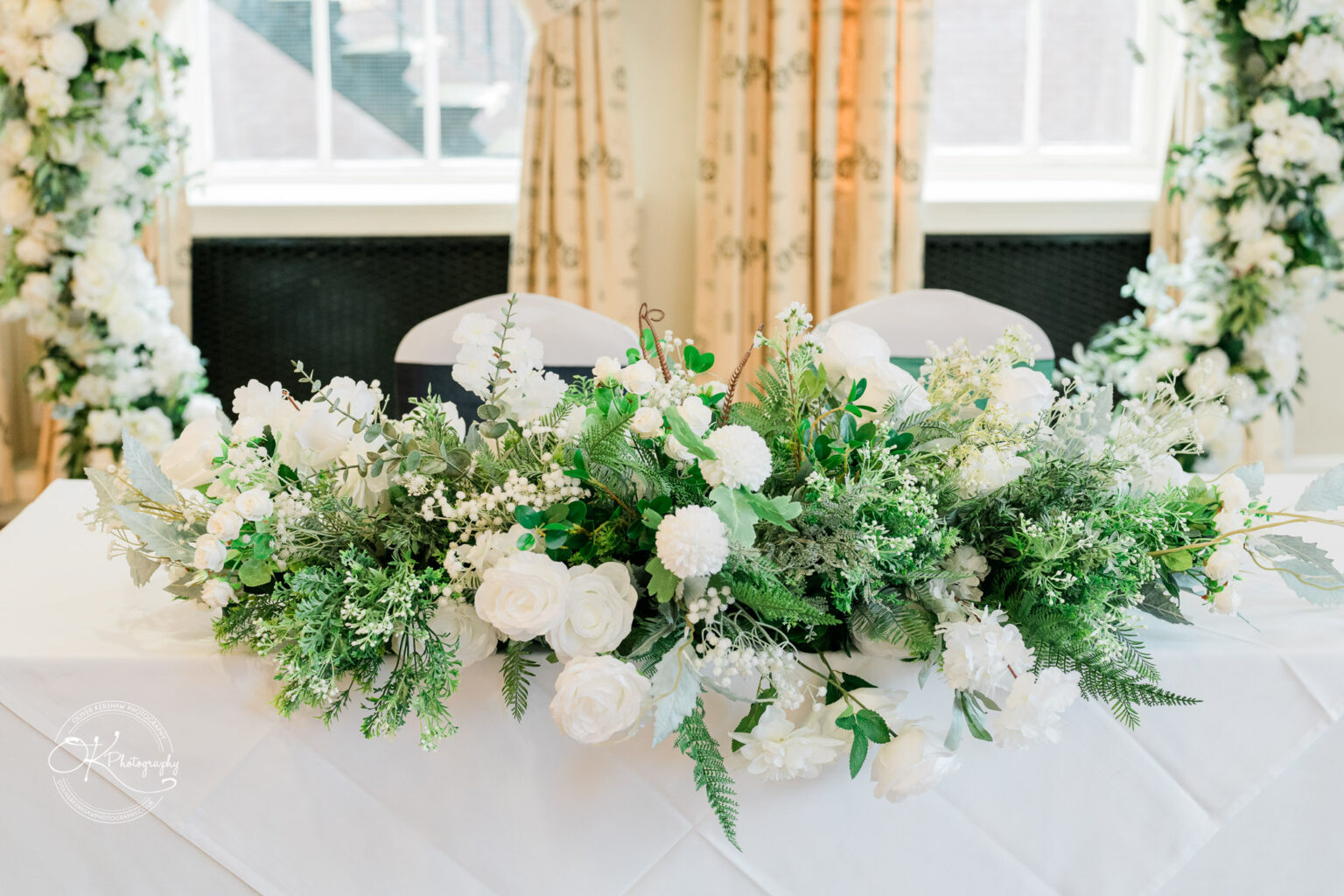 Table with a white cloth and a decorative floral arrangement of white roses and green foliage in a bright room with cream curtains and window in the background.