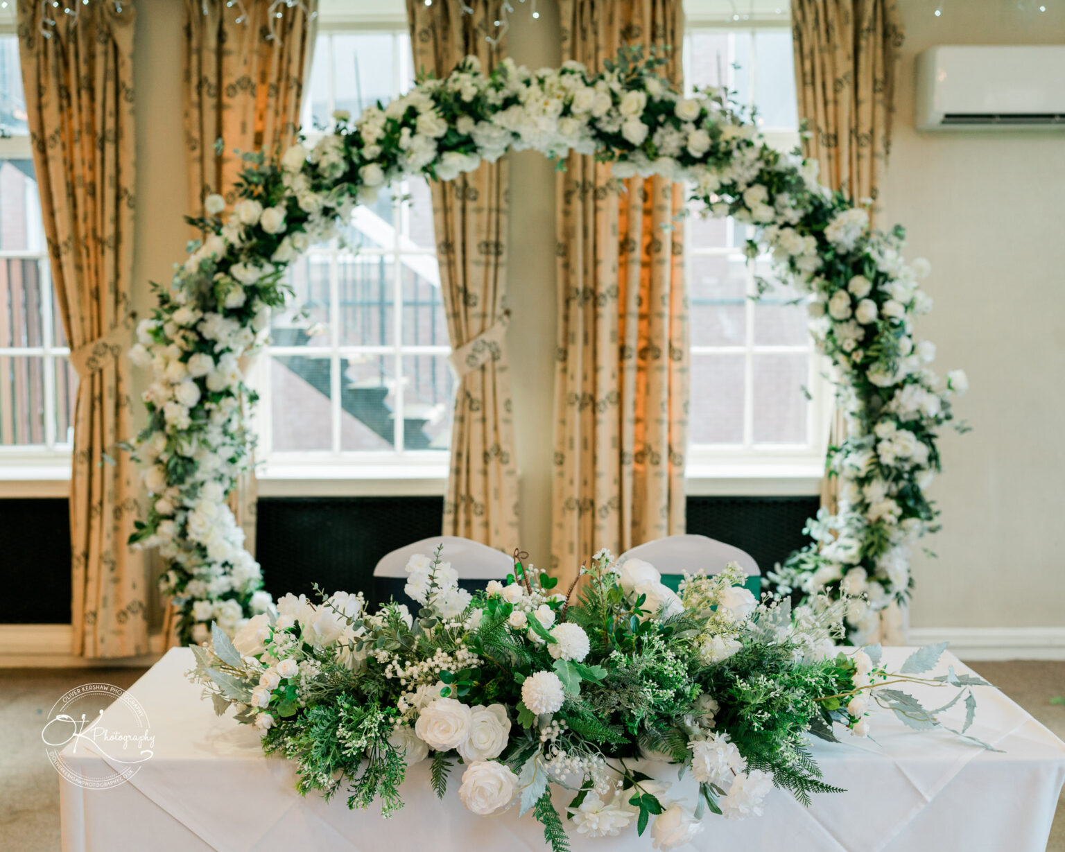 A windowed room with floral curtains featuring a white floral arch and a table decorated with green and white floral arrangements.
