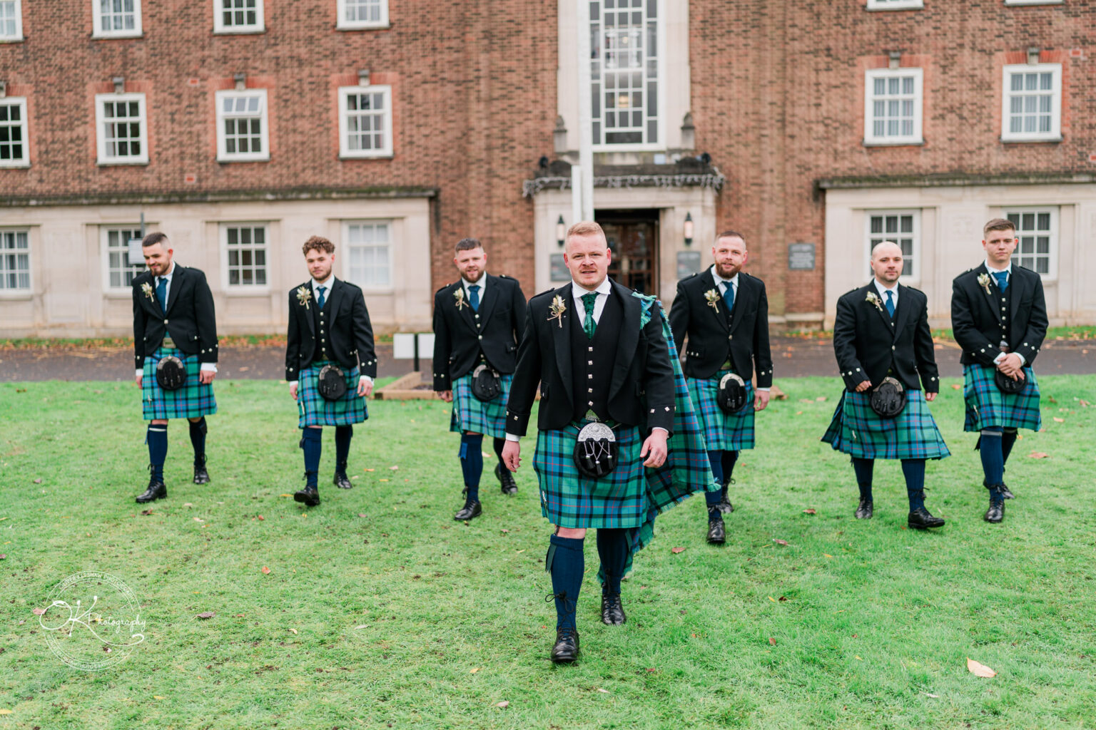 A group of men wearing traditional Scottish attire, including kilts and jackets, walking on a grassy area in front of a red-brick building with multiple white-trimmed windows.