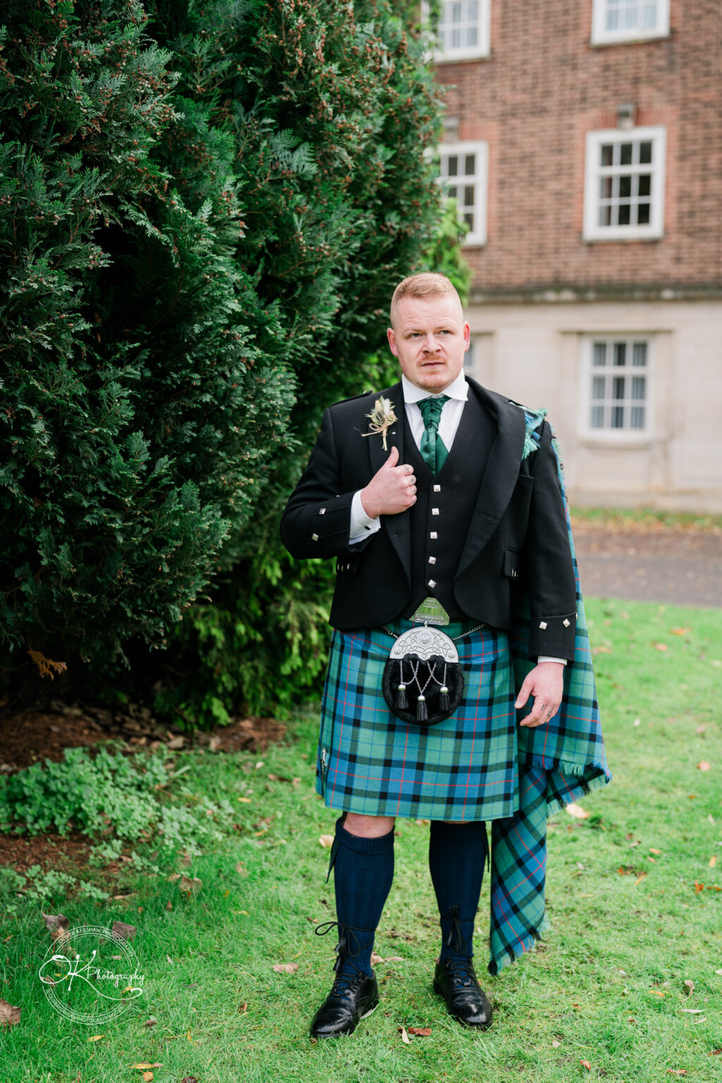 A man in traditional Scottish attire, including a kilt, jacket, and sporran, stands on a lawn beside a large tree. A brick building with white-framed windows is visible in the background.