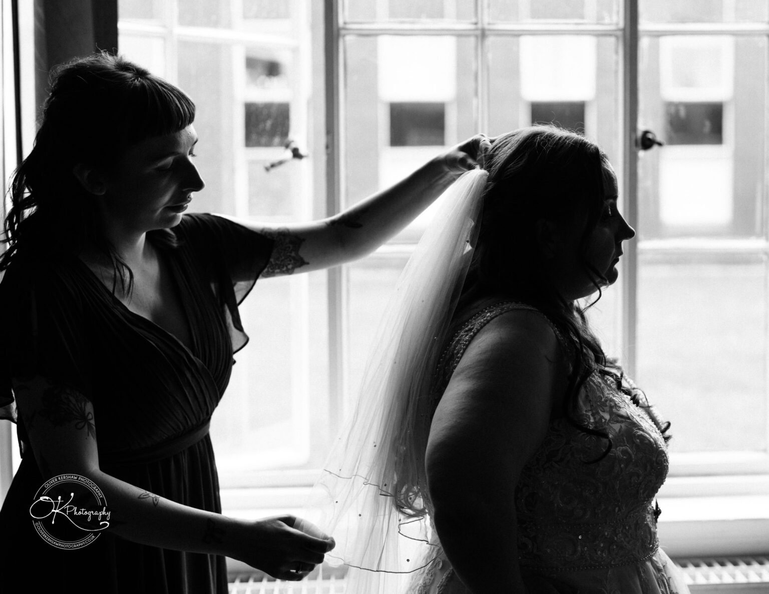 A woman helps a bride adjust her veil in front of a window, silhouettes and shadows prominent in the black and white photo.