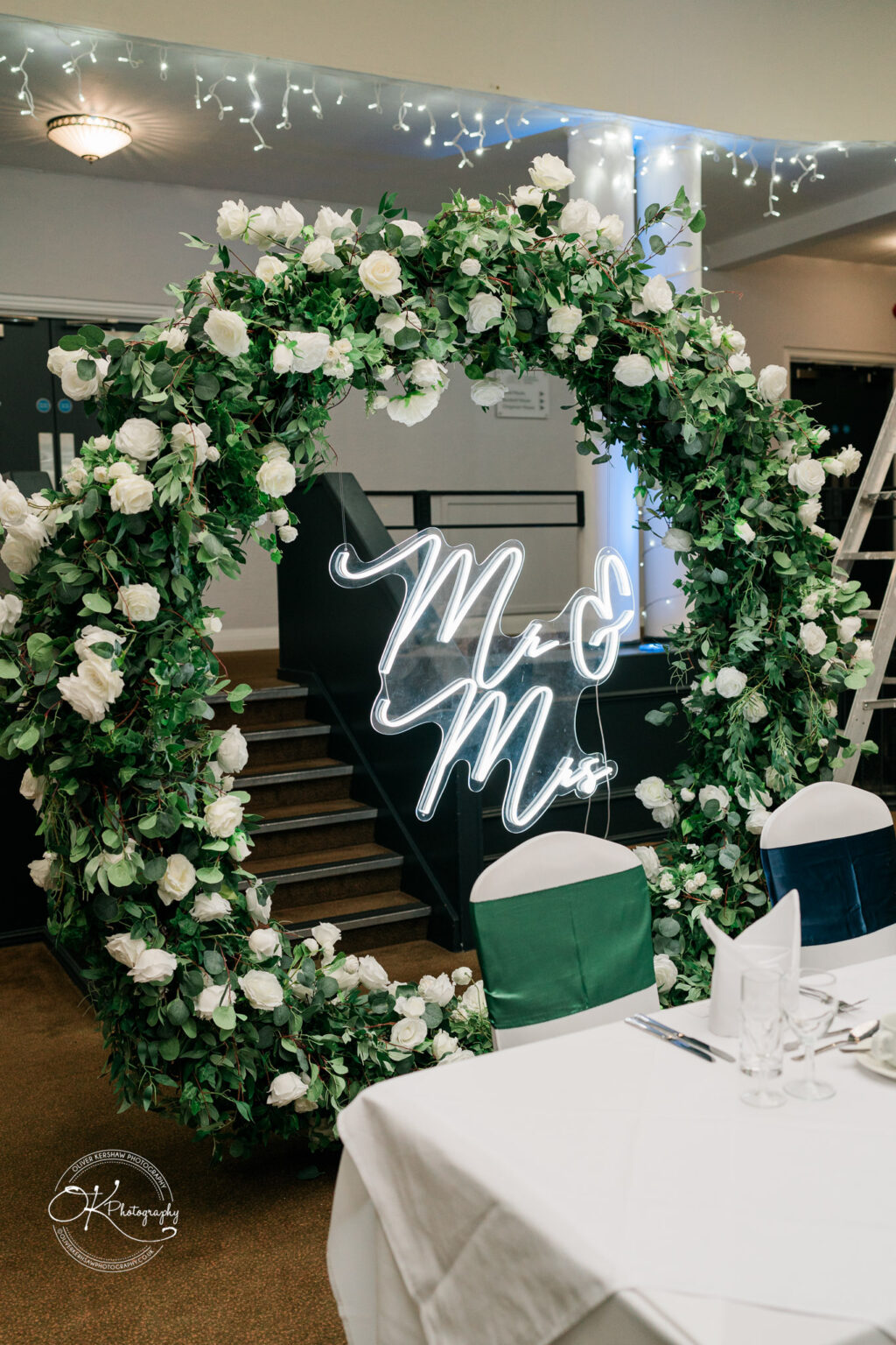 Floral arch with white roses and greenery, featuring an illuminated "Mr & Mrs" sign, at an event setup with covered chairs and a decorated table.