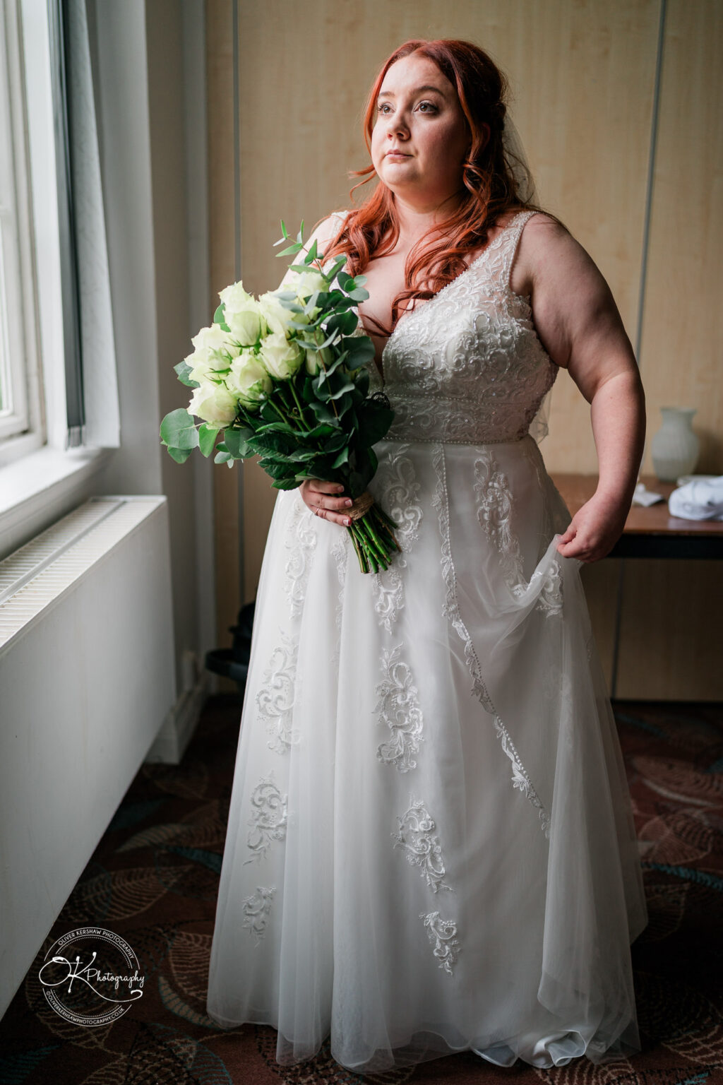 A bride in a lace wedding dress holding a bouquet of white roses stands by a window, looking thoughtfully into the distance.