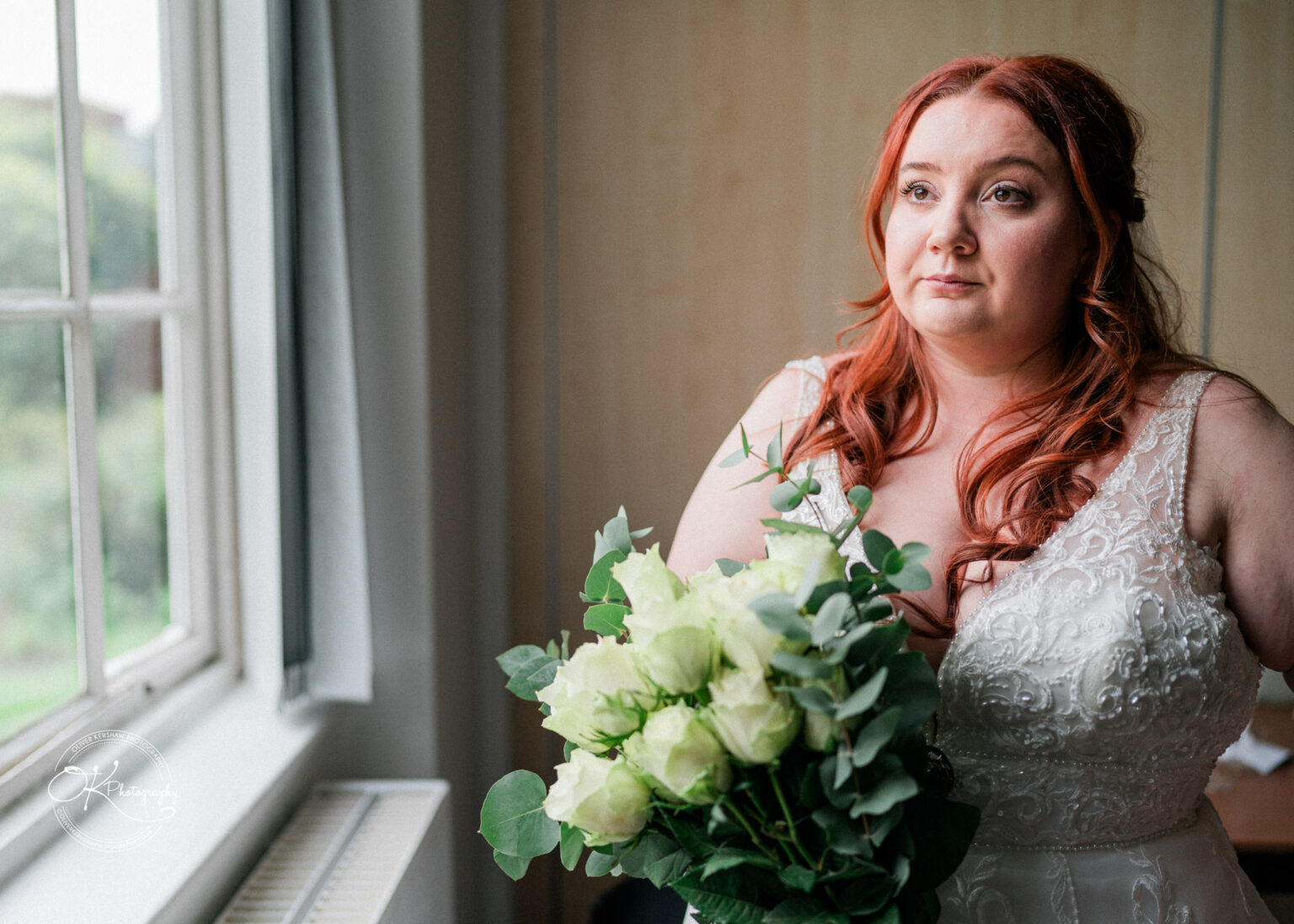 A woman in a white wedding dress holding a bouquet of white roses and greenery stands near a window.