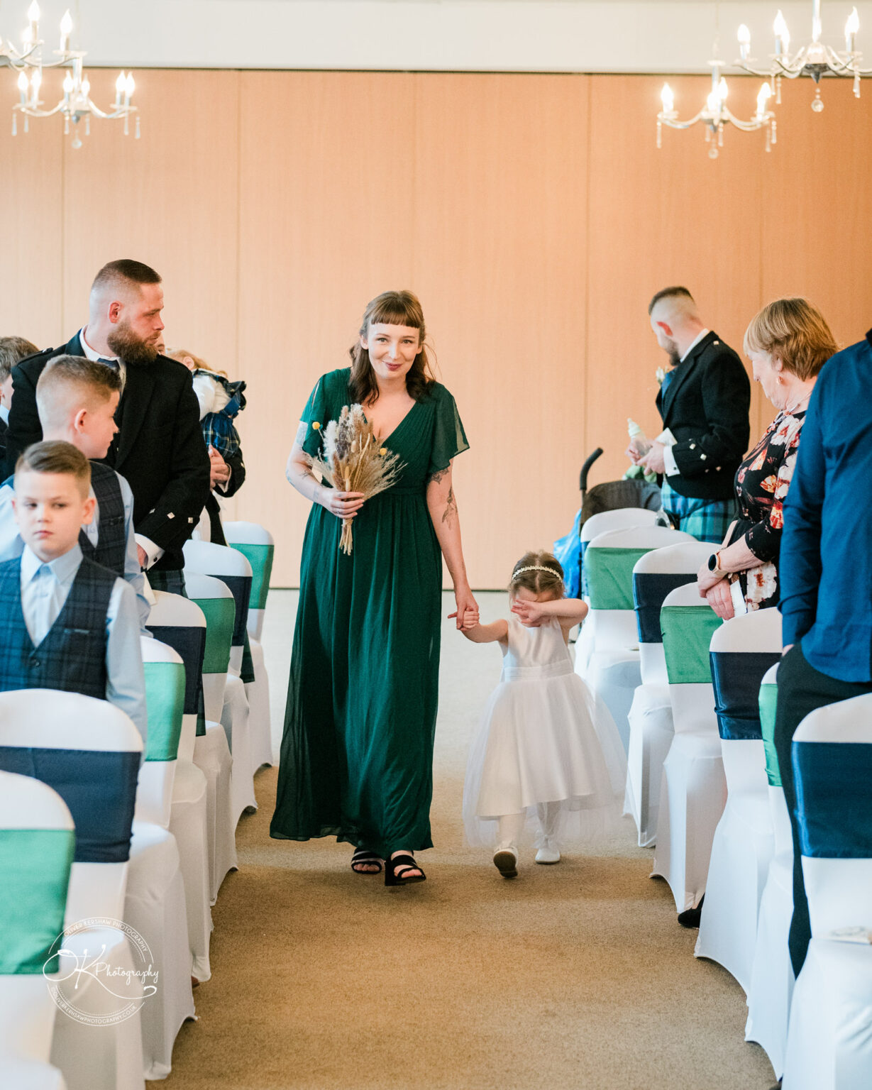 A woman in a green dress walks down an aisle holding the hand of a young girl in a white dress, who is covering her face. Guests are seated on both sides of the aisle.
