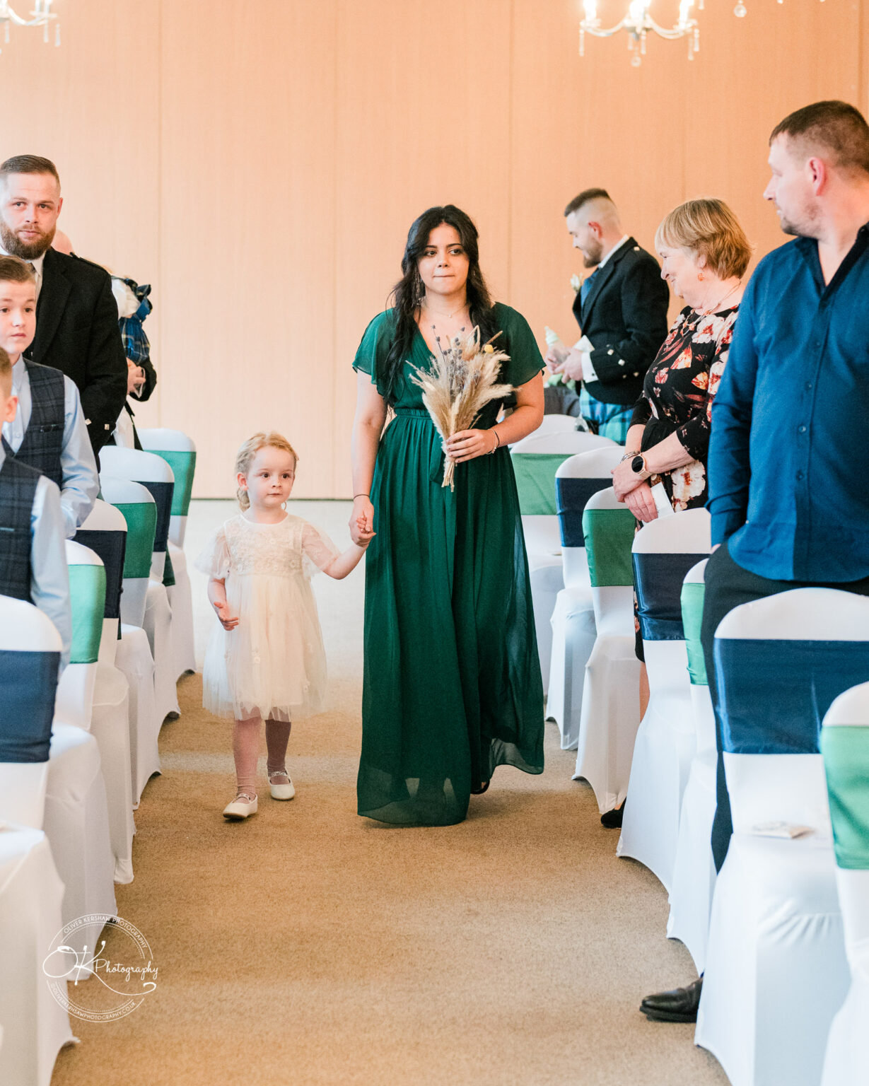 A woman in a green dress holding a bouquet and a young girl in a white dress walk down an aisle lined with chairs, while guests look on.