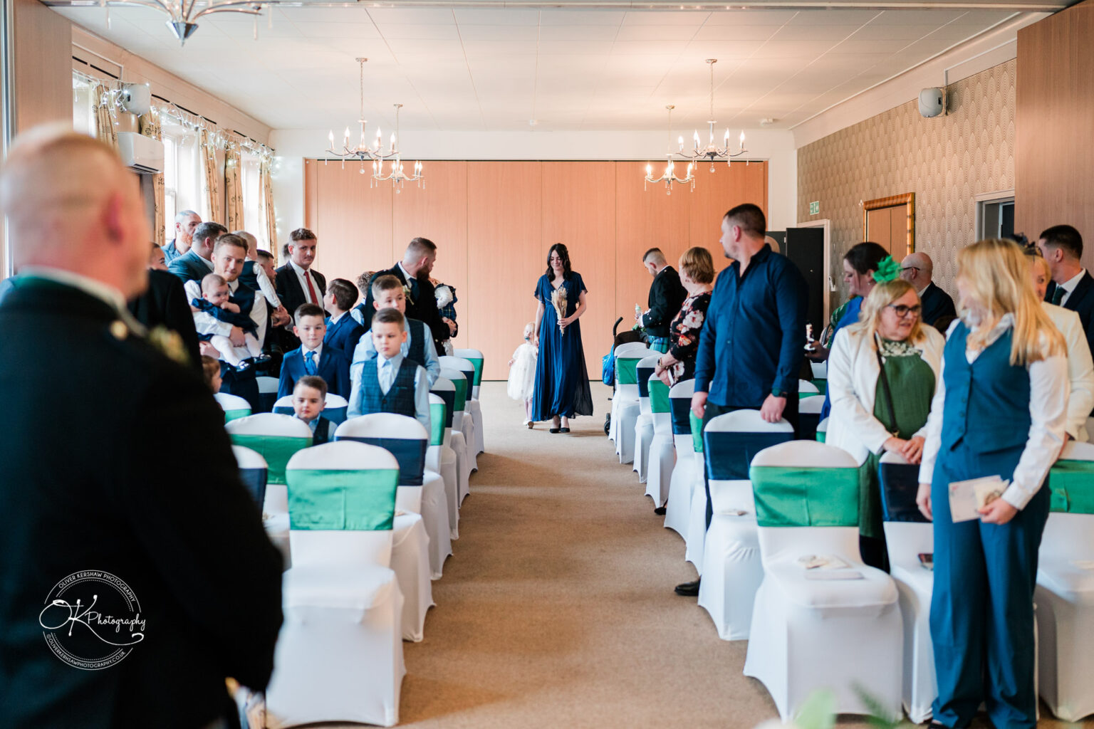 People gathering in a decorated room with white chair covers and green sashes, preparing for a ceremony at Derby Conference Centre.