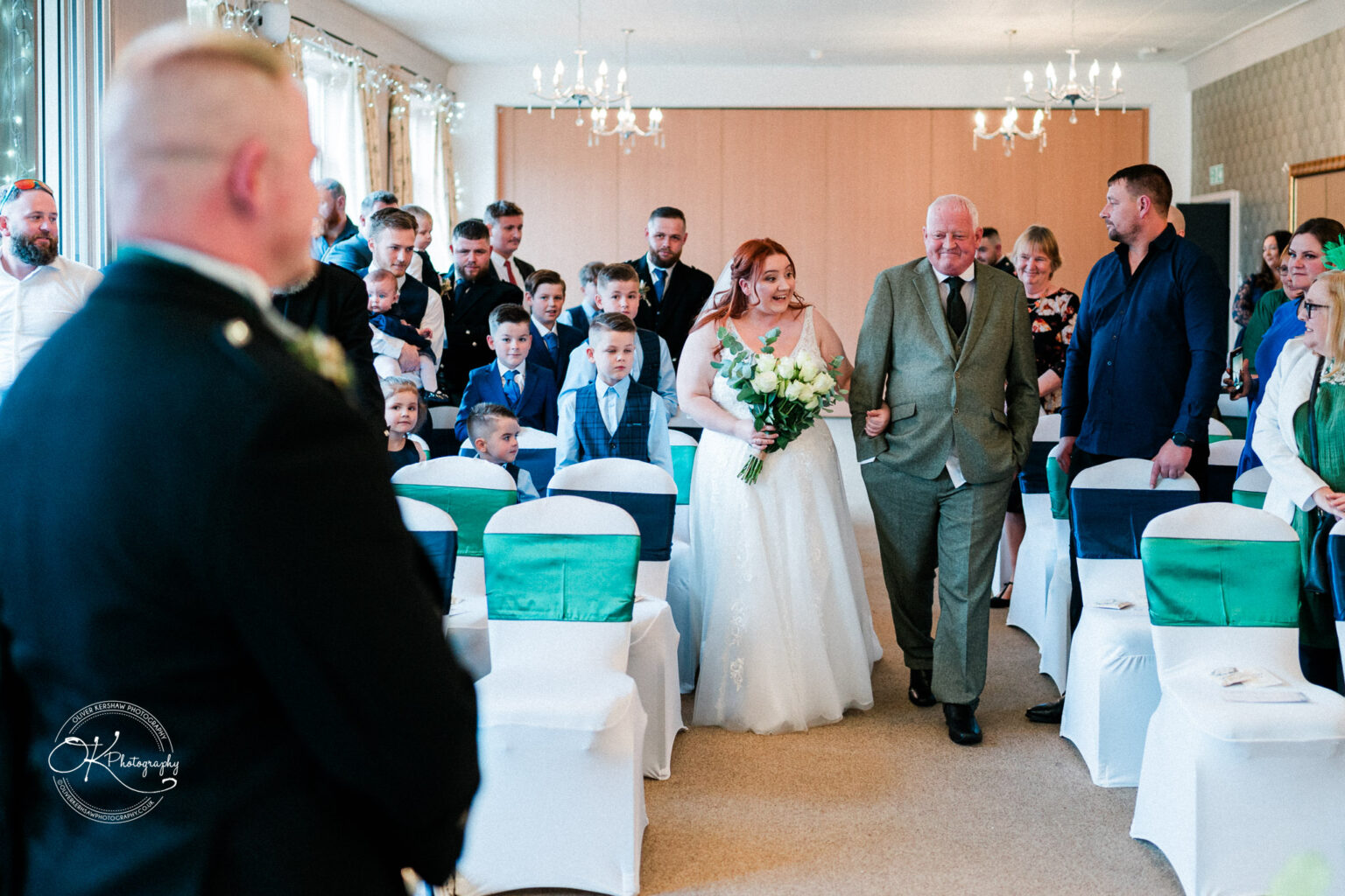Bride walking down the aisle with an older man at a wedding ceremony, guests seated and standing, some smiling and watching.