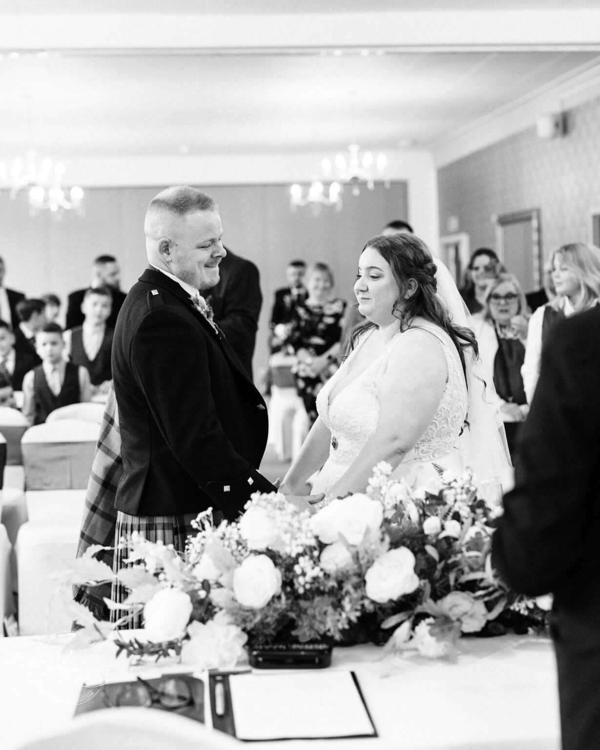 A bride and groom holding hands and smiling at each other during a wedding ceremony, with a floral arrangement in the foreground and guests seated in the background.