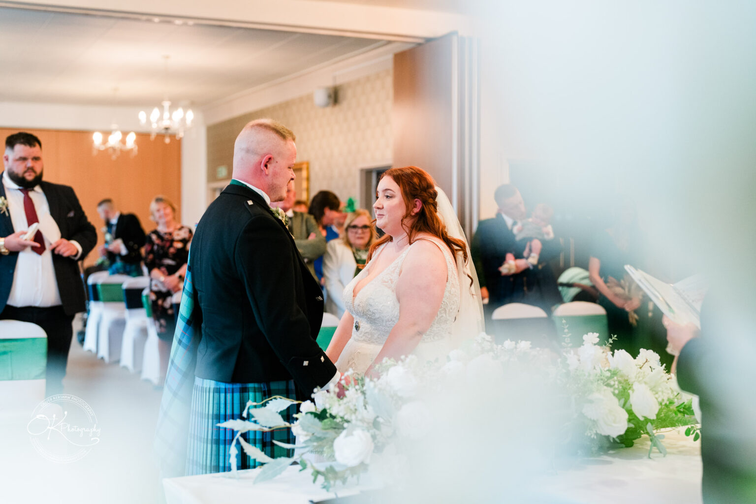 A wedding couple holding hands during their ceremony at the Derby Conference Centre, surrounded by guests.