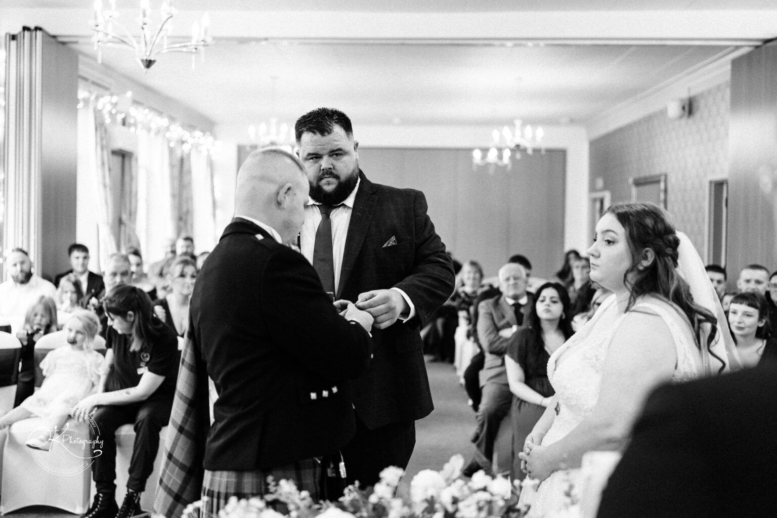 A wedding ceremony with a bride and groom at the altar, officiated by a man in a suit, in a room filled with guests.