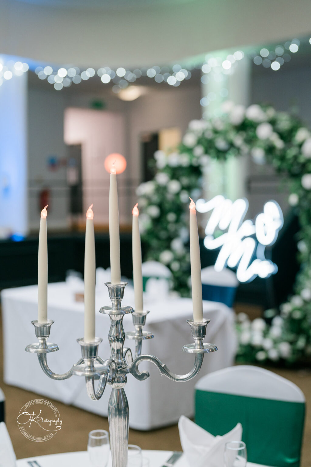 Silver candelabrum with five lit candles on a decorated table, with a floral arch and illuminated "Mr & Mrs" sign in the background.