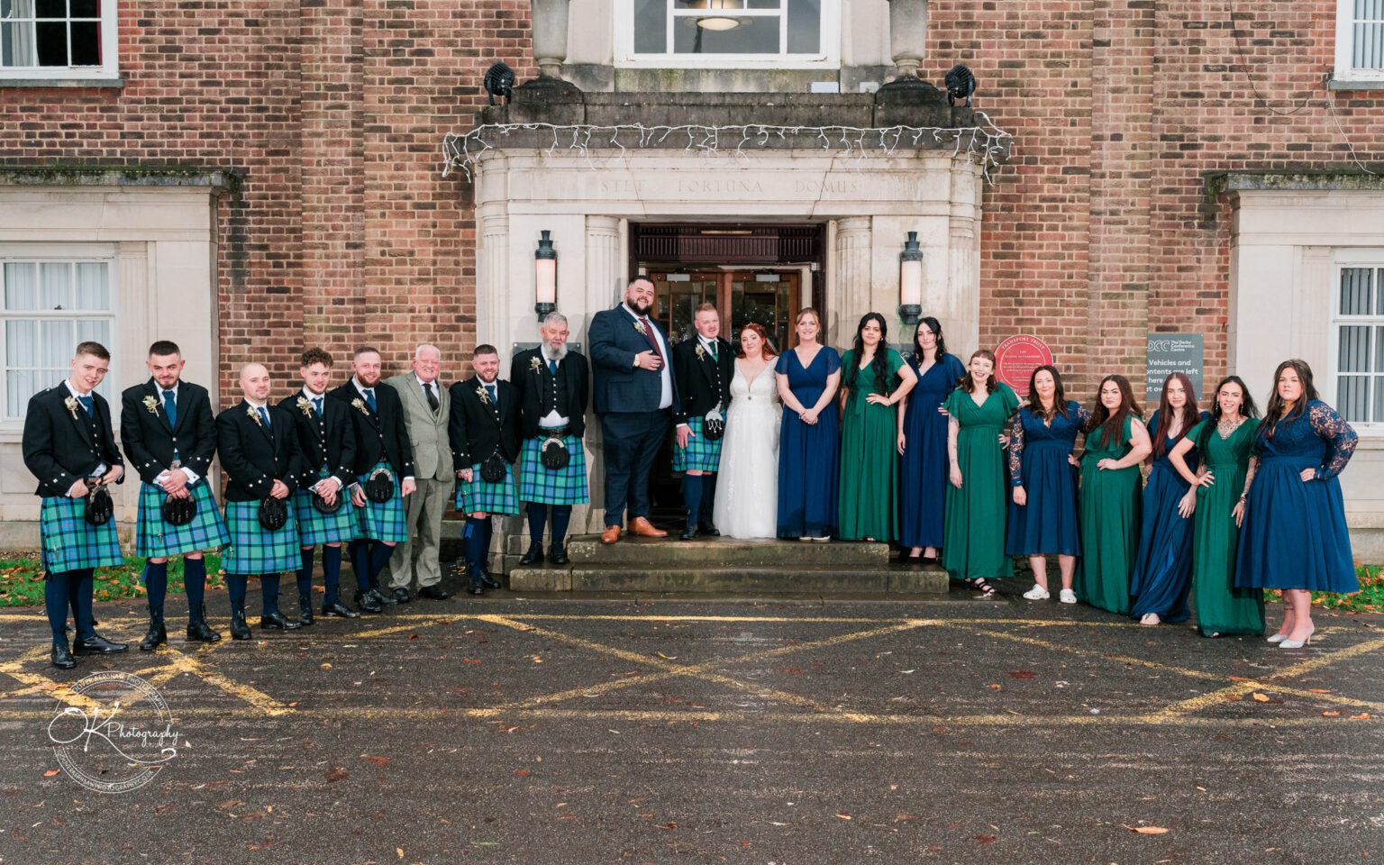 A wedding party stands in front of the Derby Conference Centre, with groomsmen in kilts on the left and bridesmaids in green and blue dresses on the right.