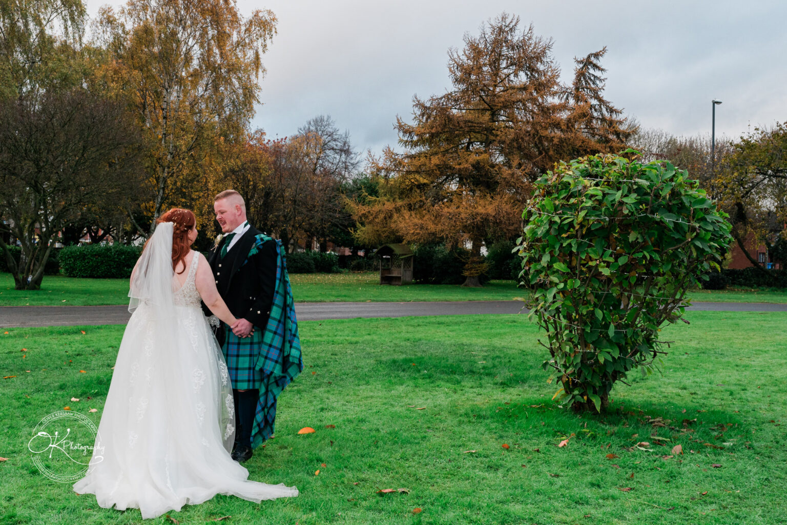 A bride in a white wedding dress and groom in traditional Scottish attire stand hand in hand on a green lawn with trees in the background.
