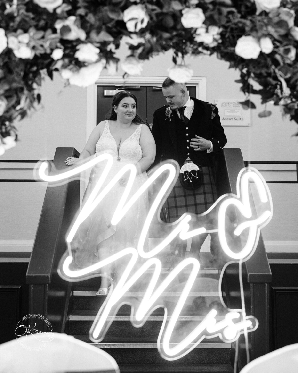 A bride and groom descend a staircase under a floral archway, with a neon sign reading "Mr & Mrs" in the foreground.