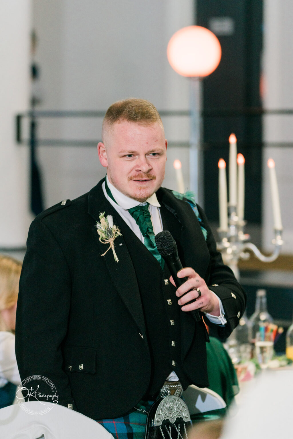 A man in traditional Scottish attire holding a microphone, possibly giving a speech, with a decorative candle stand and a blurred background.